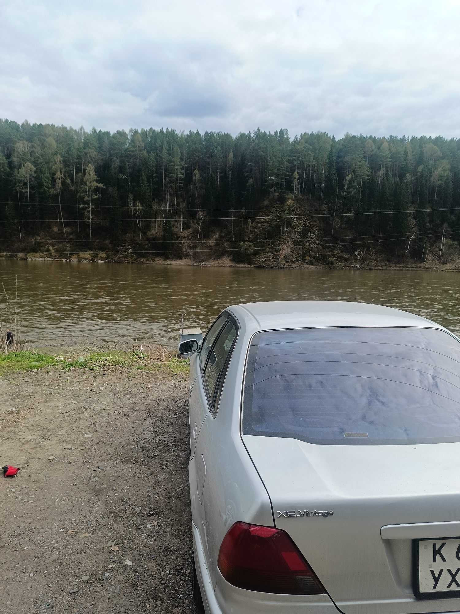 The image shows a silver "XE Vintage" car parked on a dirt and gravel bank beside a wide, brown river. Across the river is a densely wooded area. The sky is overcast. The car's license plate is partially visible. A red object lies on the ground near the car. The location appears to be a natural area outside of the city center. Based on the provided location information, the scene is likely a riverbank in or near Усть-Мана, Russia, situated along one of the region's rivers.