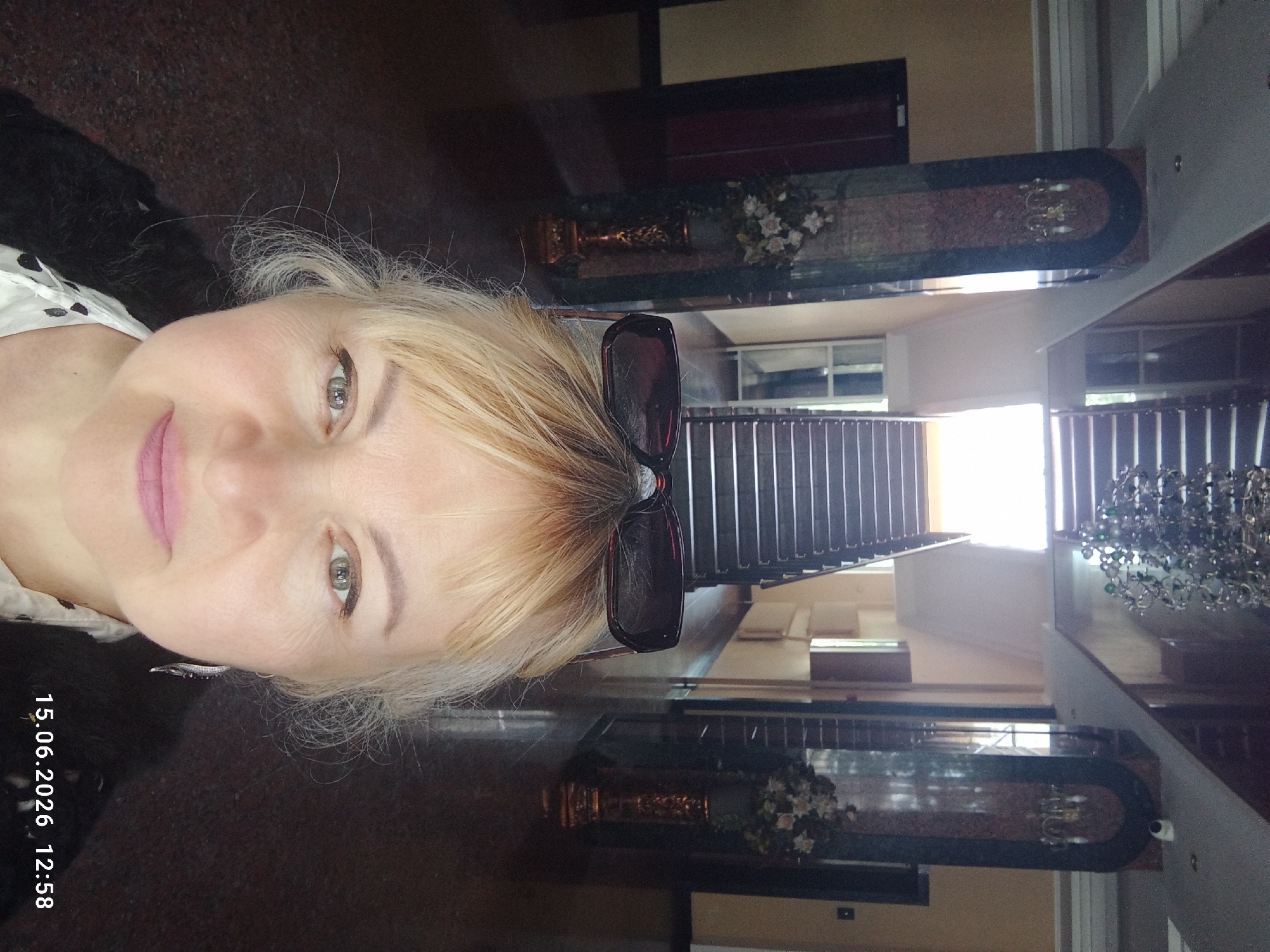 A woman is taking a selfie in what appears to be a hotel lobby or a similar indoor public space. She is positioned in the foreground, looking directly at the camera. She has blonde hair, and is wearing sunglasses on top of her head, and a black cardigan over a white patterned shirt. Her makeup is visible, with eyeliner and lipstick. She is wearing a dangling earring in her visible ear. Behind her, a grand staircase with dark treads and railings ascends to an upper level, where a large chandelier hangs from the ceiling. The walls are decorated with marble pillars and floral arrangements in ornate vases. The floor is a dark, reflective material. The lighting suggests it is daytime, with light streaming from the top of the stairs. A timestamp in the lower right corner reads "15.06.2026 12:58." The overall environment appears to be well-lit and well-maintained.
