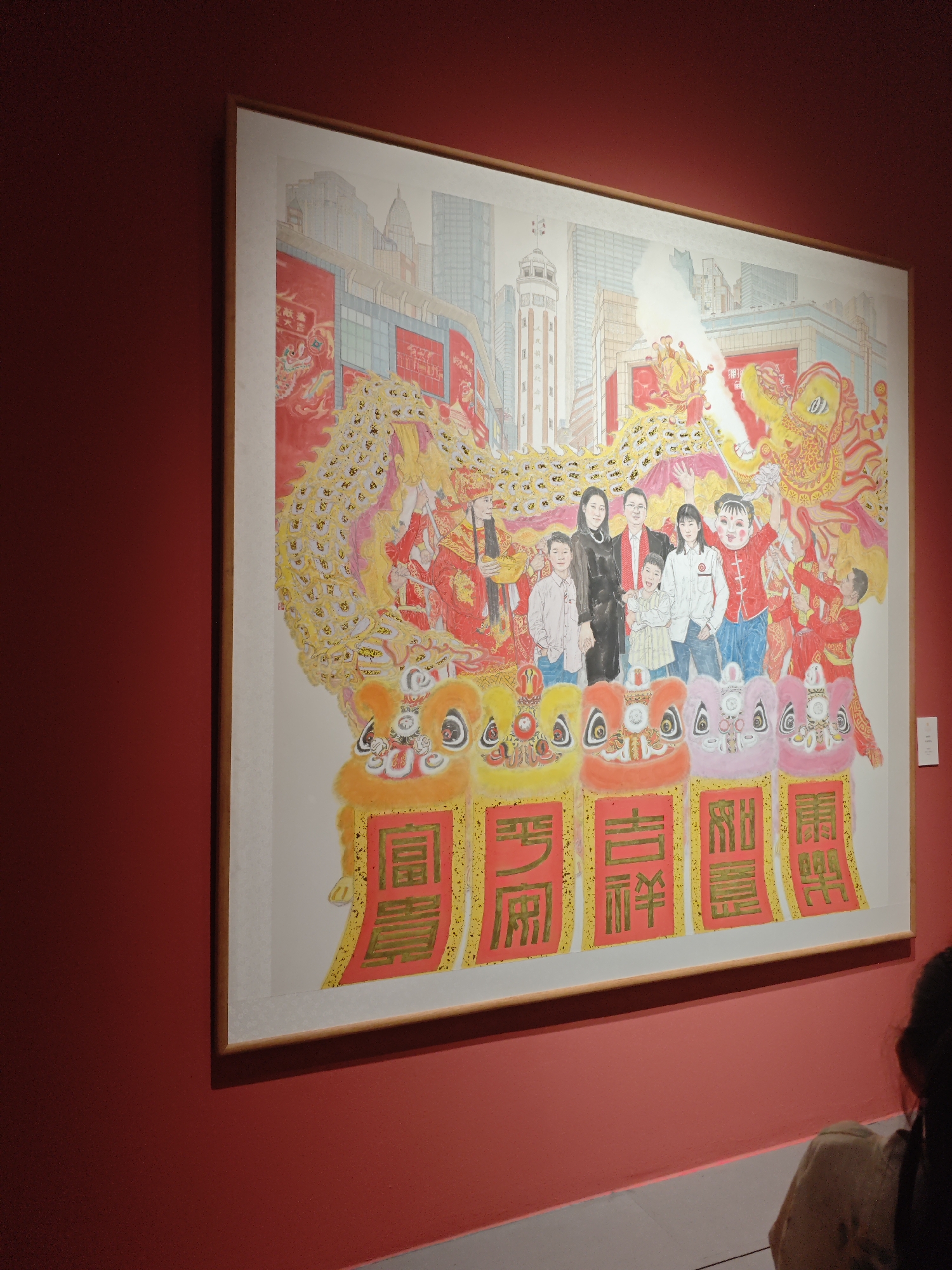 A framed painting hangs on a red wall. The painting depicts a city skyline, a family, a dragon dance, and other figures in red and gold costumes. At the bottom of the painting are four colorful lion heads, each with a gold character on a red background. A person with dark hair and a light-colored shirt is partially visible in the bottom right corner of the image. The painting is in a gallery or museum setting.