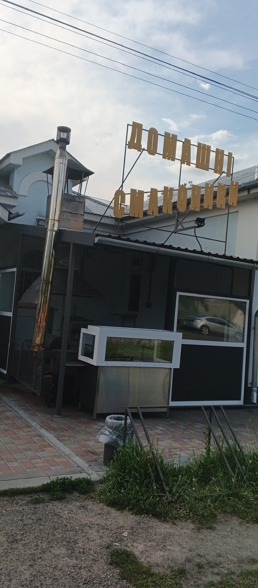 In Ploske, Ukraine, a yellow sign with Cyrillic lettering, reading "ДОМАШНІ СМАКОЛИКИ" (which translates to "homemade delicacies"), is mounted on a metal frame against a cloudy sky. Below the sign, a small open-air establishment is visible. It features a large, gleaming stainless steel chimney on the left, connected to some cooking equipment. To the right, there are large display windows, one of which reflects a silver car. The area in front of the establishment is paved with brick-like tiles and gravel. In the foreground, there is a trash can, some green grass, and a metal rack.