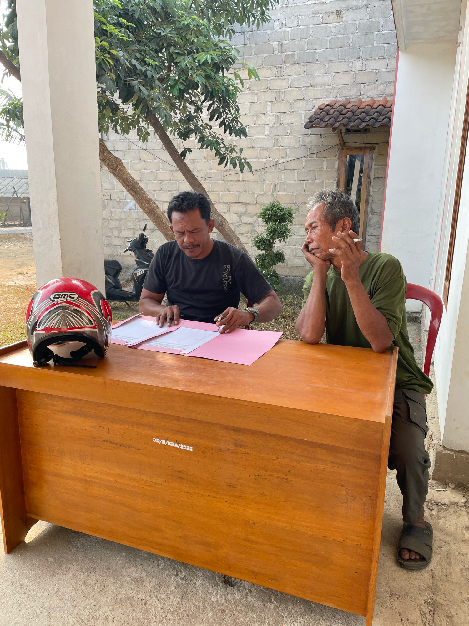 In the city of Kagungan Ratu, Indonesia, two men are seated at a wooden desk outdoors, positioned near a white brick building. The man on the left, wearing a black t-shirt, is focused on papers spread across the desk, with a pen in his hand. A red and silver motorcycle helmet rests to his left. The man on the right, dressed in a green t-shirt and dark pants, leans his head on his hand and holds a lit cigarette. A red plastic chair is behind him. The environment is somewhat rural, with trees and a dirt ground visible. The desk has some markings on its front, which appear to be "DD/II/KRA/2024".