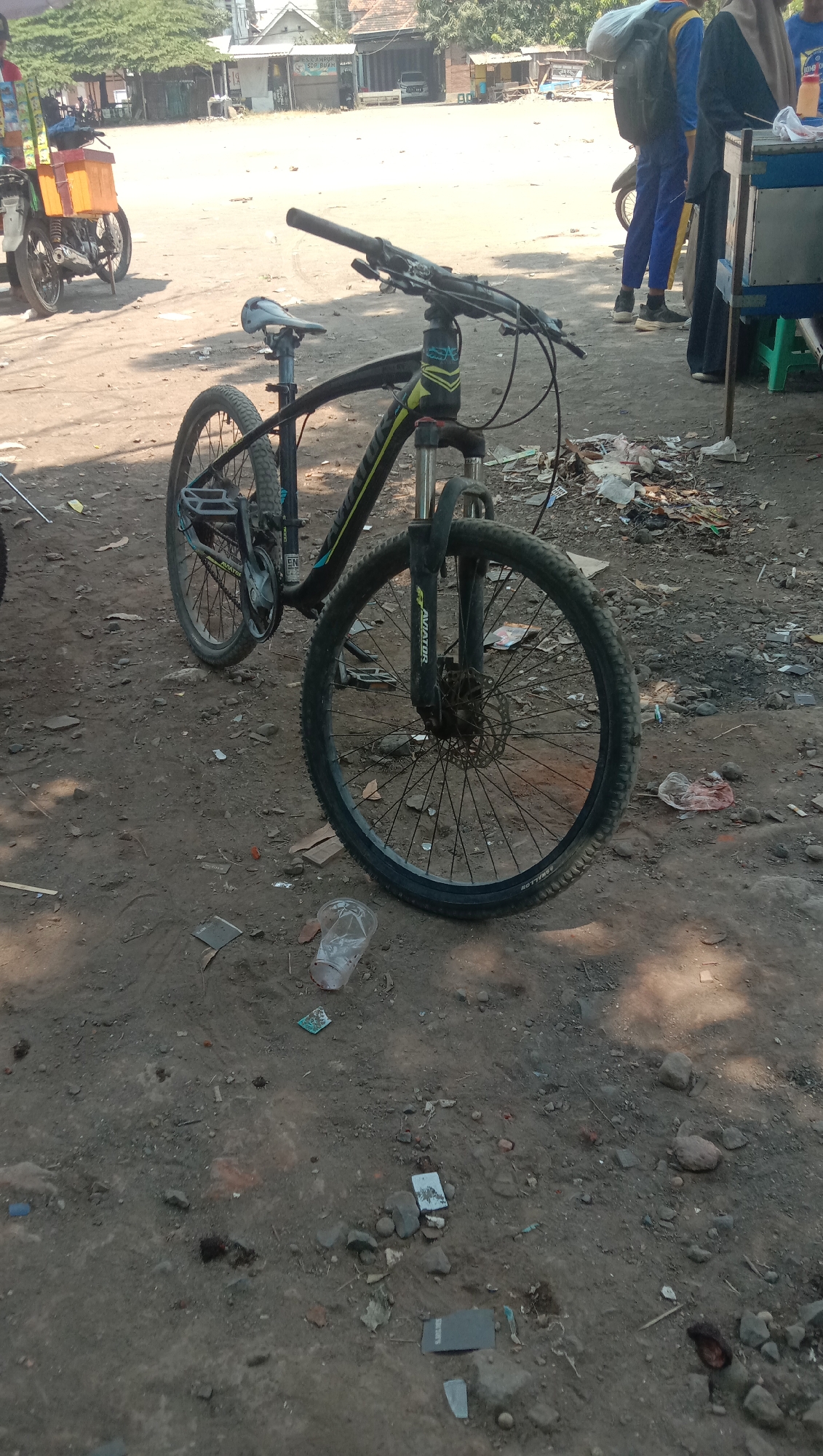 A bicycle is parked on a dirt road in what appears to be a commercial or roadside area. The bicycle is black and yellow, with a front suspension fork and disc brakes. It has a black seat and pedals. In the background, there are some buildings with awnings, suggesting a marketplace or shops. To the left, a motorcycle with an orange cargo box is visible. Further in the background, several people are walking, some carrying bags. The ground is uneven and covered in dirt and small rocks, with scattered debris including plastic cups and wrappers. The overall impression is of a busy, informal outdoor setting.