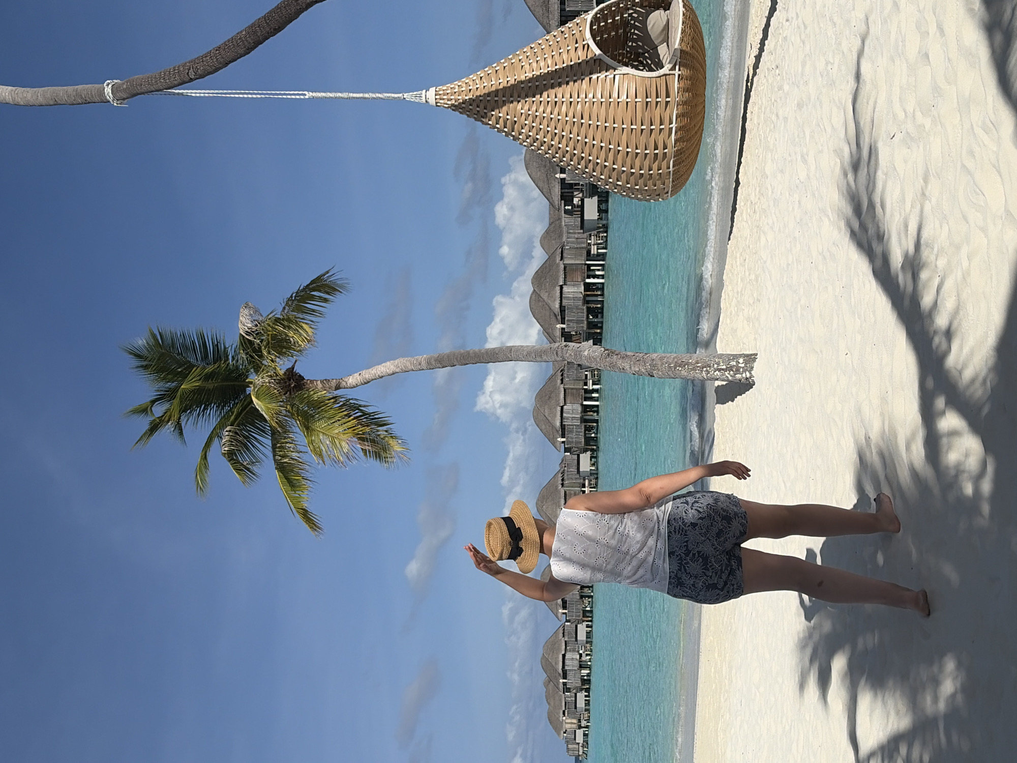 A woman wearing a straw hat and a white top with a floral pattern walks on a white sandy beach in Halaveli, Maldives. She is facing away from the camera, looking towards a row of overwater bungalows in the turquoise ocean. To her right, a large woven hanging chair is suspended from a palm tree. Palm tree shadows are cast across the sand. The sky is blue with scattered clouds.