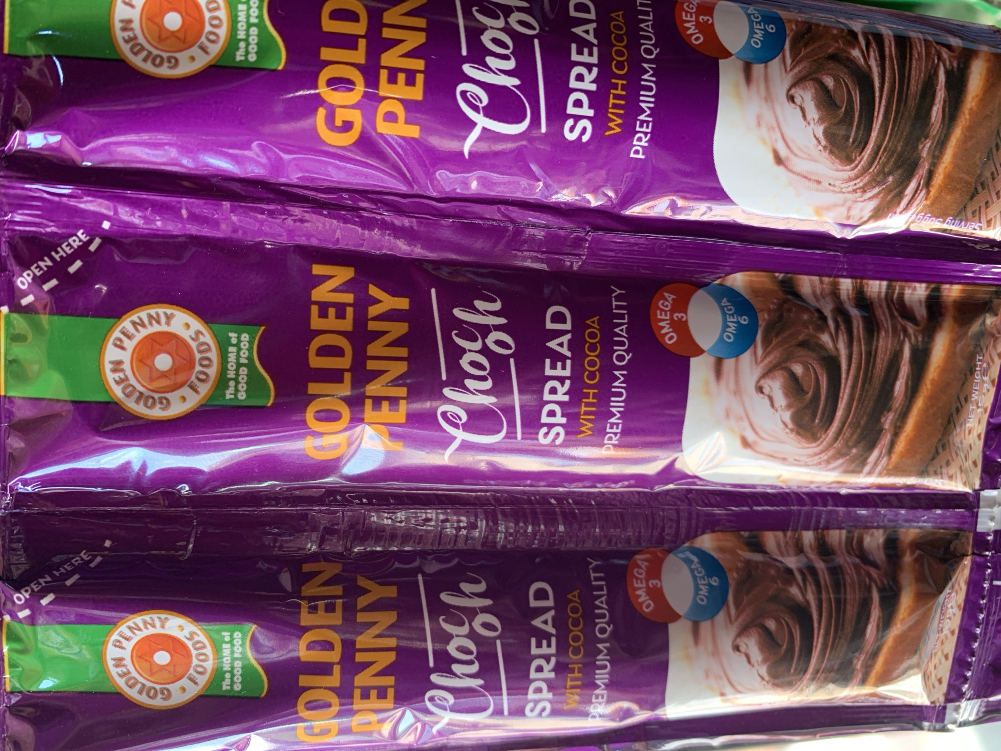 The image shows three packages of Golden Penny Choc Spread arranged in a vertical stack. The packaging is purple and displays the brand name "GOLDEN PENNY" in orange text, along with "Choc SPREAD WITH COCOA PREMIUM QUALITY" in white text. There is an image of the spread on a piece of bread and labels "OMEGA 3" and "OMEGA 6". The top of the package indicates "OPEN HERE" with dashed lines. No individuals or specific environmental details suggesting a location in Rafin Par, Nigeria, are discernible in the image.