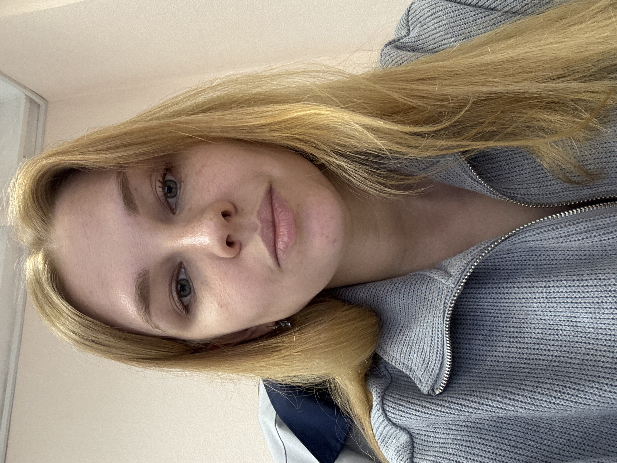 A close-up, rotated image shows a fair-skinned person with long, light blonde hair. The person is wearing a gray zippered sweater or jacket. The zipper is visible. The background is a light neutral color. The person is looking directly at the camera with a neutral expression. Due to the lack of visible environmental details, it is impossible to determine the location as Rusanovo, Russia. There are no actions, interactions or specific objects other than clothing. The scene is confined to the person's head and upper torso.