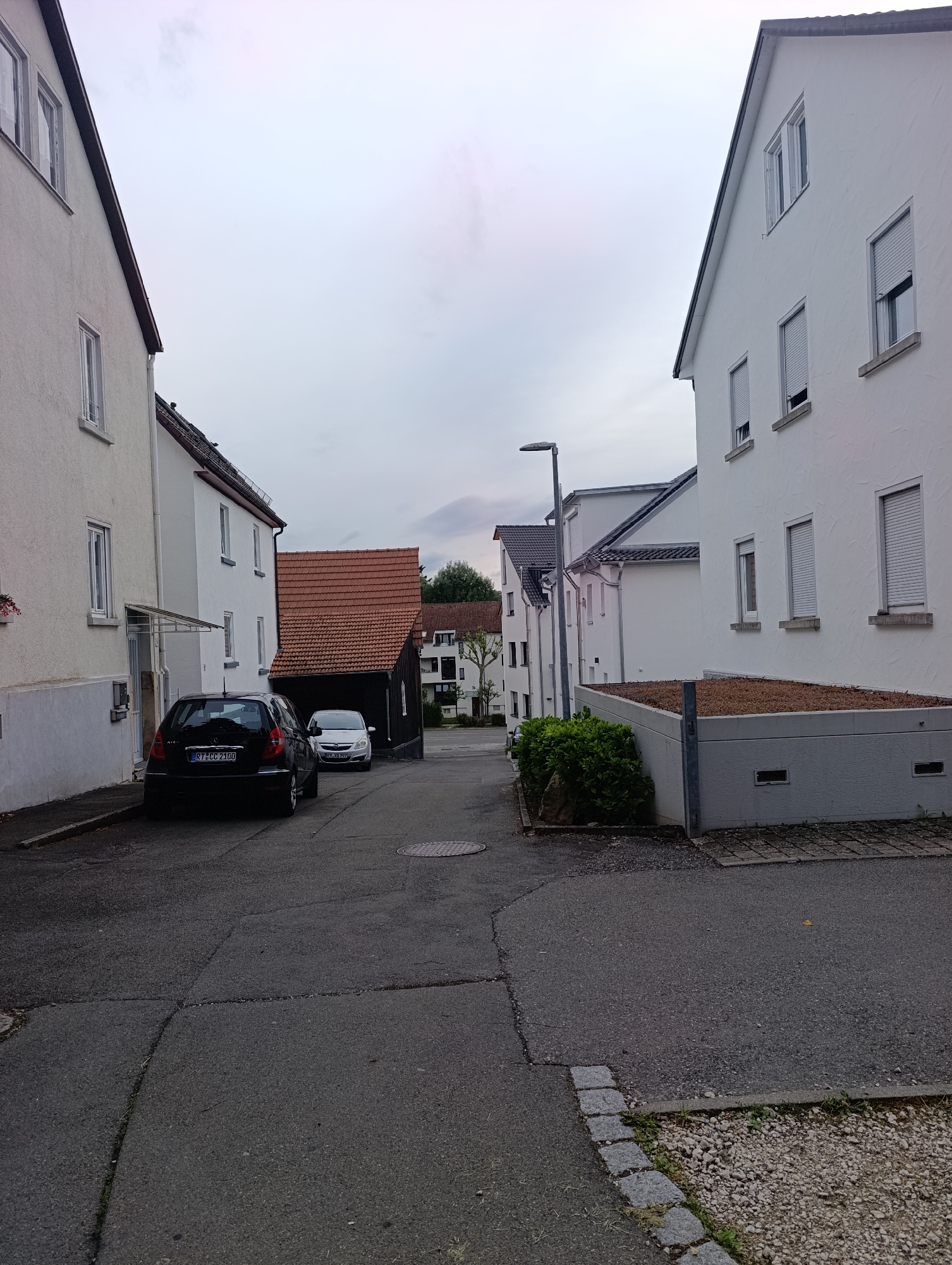 The image depicts a street scene in Pfullingen, Germany. On the left, a white residential building with a gray base and several windows is visible. In front of it, a black hatchback car is parked, displaying a license plate that reads "RT CC 2100." Further down the street, a white compact car is parked. Adjacent to the black car is a dark wooden structure with a red tiled roof.
On the right side of the street, a modern white residential building with multiple windows, some equipped with roller shutters, is prominent. A low, gray concrete structure with a gravel-covered top and ventilation openings is situated in front of this building. A small patch of green bushes is located to the left of this structure, and a large rock is partially visible within the bushes. A street lamp stands further down the street. The road surface is asphalt, marked with various cracks and a manhole cover. The sky is overcast.
