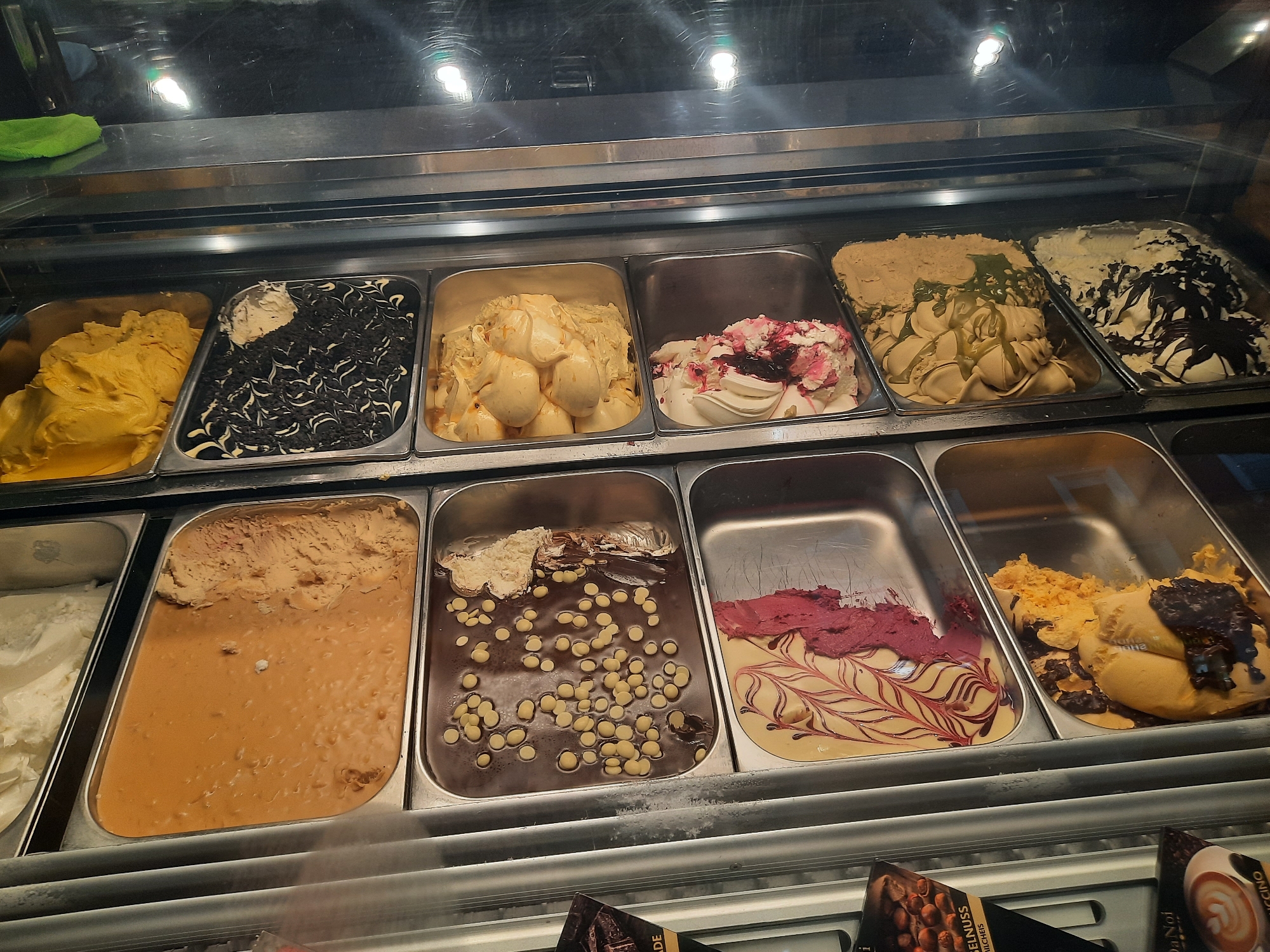 This is a medium shot, eye-level, indoor photograph of an ice cream display case in a shop, likely a gelato or ice cream parlor. The display case contains multiple metal trays filled with various flavors of ice cream.
From left to right, the top row shows:
1. A tray filled with bright yellow ice cream.
2. A tray with dark chocolate ice cream swirled with white cream and what appear to be cookie crumbles.
3. A tray of pale yellow ice cream with visible chunks and swirls, possibly caramel or vanilla bean.
4. A tray of white ice cream with red fruit compote or swirls.
5. A tray of light brown ice cream with a greenish drizzle and some white chunks.
6. A tray of pale yellow ice cream with dark chocolate drizzles and chocolate chips.
The bottom row appears to contain:
1. A tray of white ice cream.
2. A tray of light brown, creamy ice cream with visible peanut or nut pieces, covered in a thin, liquid caramel-like sauce.
3. A tray of dark chocolate ice cream studded with numerous white chocolate chips or candies.
4. A tray of pale yellow ice cream with red fruit swirls and dark chocolate drizzles.
5. A tray of yellow-orange ice cream with dark chocolate pieces and a dark, syrupy topping.
The environment is brightly lit, reflecting off the glass of the display case. A green cleaning cloth is visible on the counter above the display. The overall impression is a clean, well-stocked ice cream shop. There are no people visible in the frame. The photograph is taken in a shop located in Rosenheim, Germany, as indicated by the context provided.