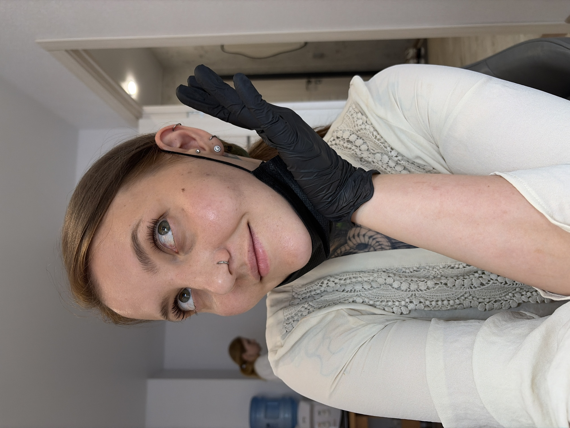 The image shows a medium shot of a young Caucasian woman with light brown hair and green eyes, wearing a light-colored blouse with lace detailing. She has a nose ring and ear piercings. A black disposable mask is pulled down below her chin, and she is wearing a black glove on her right hand, which is positioned near her face as if she is posing. She is looking up and to her left, with a slight smile. In the background, a water dispenser is visible, and another person, also a woman with blonde hair, is partially seen in the distance. The environment appears to be an indoor space, possibly an office or a clinic, with a doorway and a light fixture visible in the background. The image does not contain any specific markers that would allow for definitive identification of the location as Izhevsk, Russia.