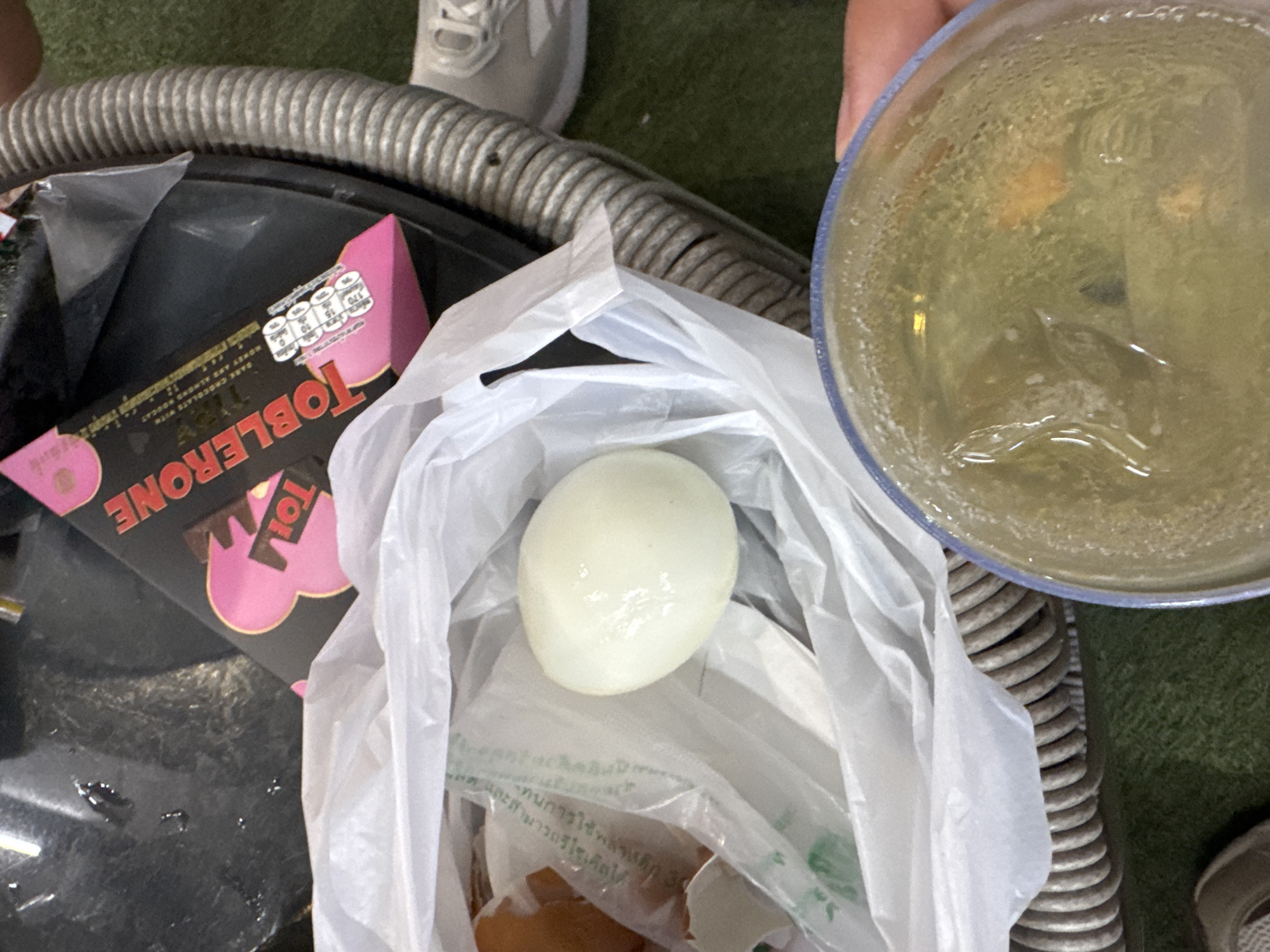 The image shows a close-up view of a boiled egg, partially peeled and resting in a plastic bag. Next to the bag is a partially opened Toblerone chocolate bar. In the upper right corner, a glass or bowl contains a yellow liquid, possibly a drink or a mixture of egg whites and yolks. The subjects are a boiled egg and Toblerone. The scene appears to be outdoors, possibly on a grassy surface, with a ribbed hose-like object visible in the background. There are no clear indications of time of day or weather. The overall impression is casual, possibly a snack break.
Visible text: The Toblerone bar has "TOBLERONE" written prominently on its packaging. There is also some Thai text on the plastic bag that holds the egg.