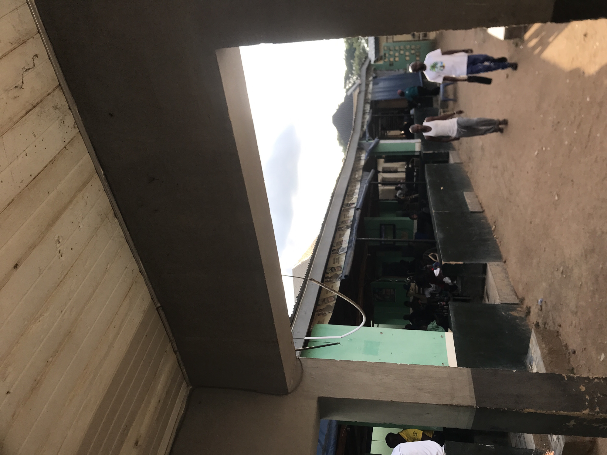 The scene is an unpaved outdoor area in New Stadium, Nigeria, viewed from beneath a concrete overhang. The ceiling of the overhang consists of light-colored, stained wooden planks. A thick, dark grey concrete beam spans the upper frame, supported on the left by a two-toned concrete pillar, grey at the top and darker, stained grey at the base. From the overhang, a black wire, a white curved tube, and a light-colored rope hang down.
To the left of the dirt path, a row of low-rise structures with light green walls and black bases features open storefronts and corrugated metal roofs. Banners or posters are affixed along the fascia beneath the roofs. Inside these establishments, multiple individuals are visible, some seated. In the immediate foreground on the left, parts of two individuals, one in a white shirt and another in a yellow shirt, are visible behind the pillar.
The ground is a reddish-brown dirt path, extending into the distance. Two adult males walk away from the viewer along this path. The man on the left wears a white sleeveless shirt and grey trousers. The man on the right wears a white t-shirt with a graphic design, blue jeans, and carries a black bag in his left hand. Strong shadows are cast on the path towards the right, indicating the direction of sunlight.
In the background, additional structures are visible, including one with a dark, conical roof. Beyond these buildings, trees or dense foliage are present. The sky above is bright and partly cloudy. On the far right, a light-colored fence or gate with a patterned design is partially visible.