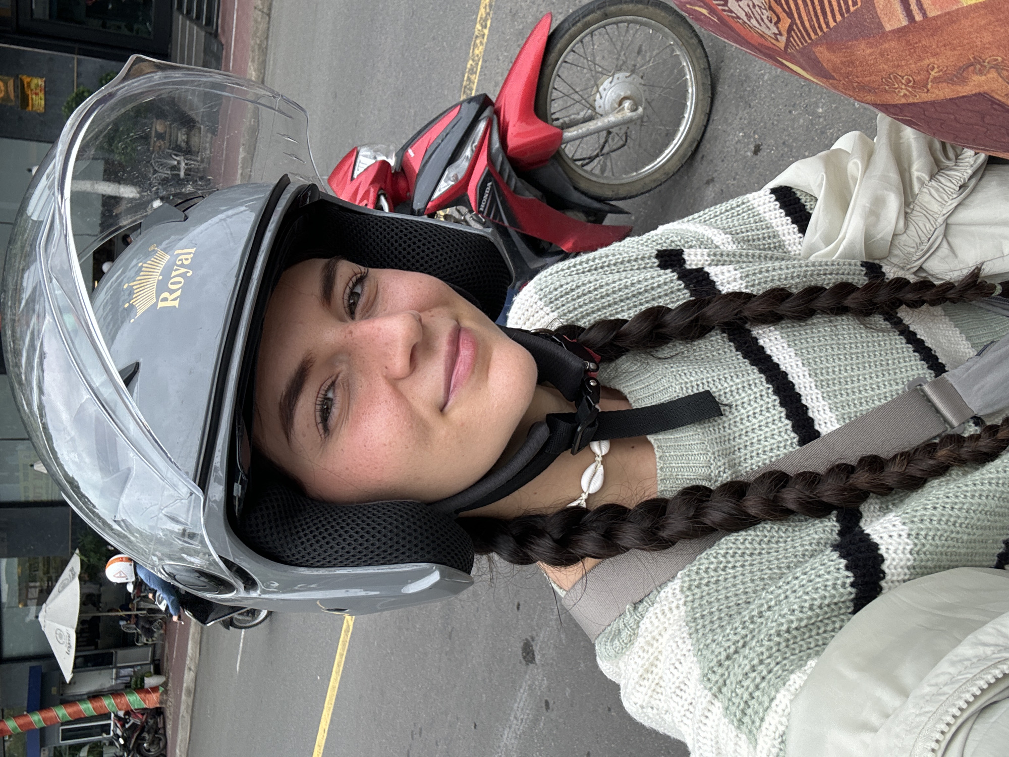 A woman is wearing a gray "Royal" motorcycle helmet with a clear visor and a black buckle. She has dark hair styled in braids. She is wearing a light green and black striped sweater and a shell necklace. A red and black motorcycle is visible in the background. The woman appears to be in Phan Thiết, Vietnam, based on the tropical foliage and street scene in the background. The street has a yellow line marking. Other parked motorcycles and storefronts are visible in the background.