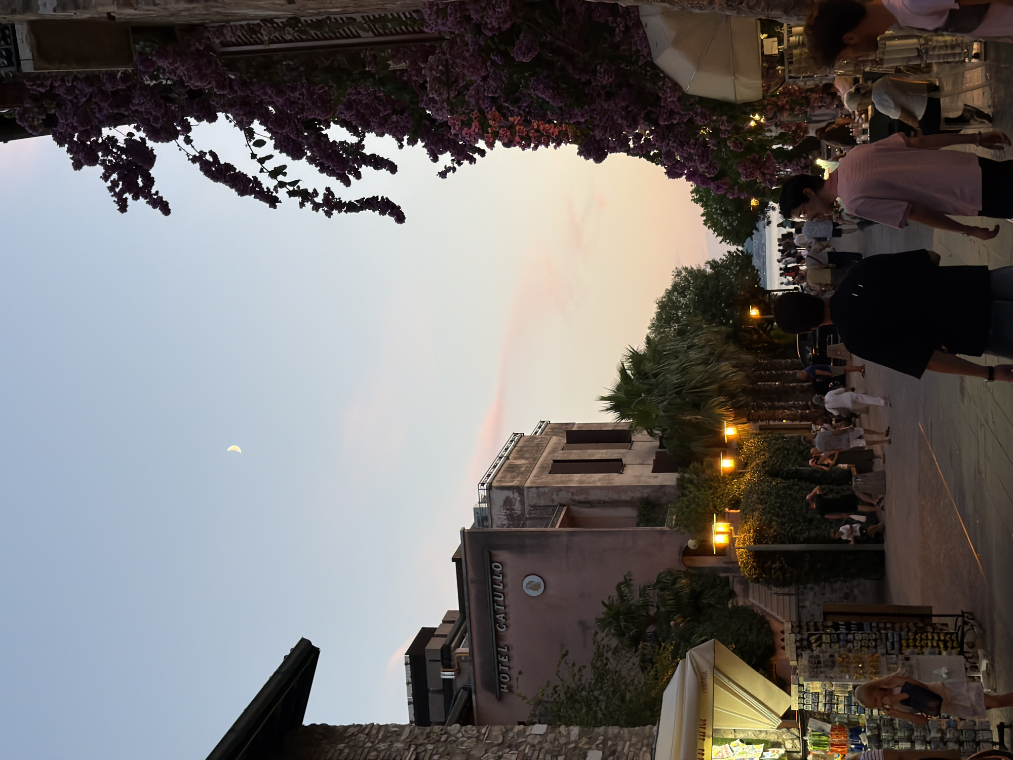 The image depicts a street scene in Sirmione, Italy, at dusk. A crescent moon is visible in the pale blue sky, tinged with pink hues from the setting sun. On the left, a building with stone accents and a dark roof is visible. To its right, a light pink building with the inscription "HOTEL CATULLO" stands out, featuring dark shutters and a circular emblem. Lush green palm trees and dense foliage line the street. Vibrant purple bougainvillea cascades down the right side of the frame, obscuring a portion of the sky and a building.
The street is paved and illuminated by warm yellow streetlights. Several people are present: a young blonde girl in a white dress stands near a display of souvenirs on the left. Further down the street, a group of adults are walking and socializing. Two young men in dark clothing are walking towards the viewer in the foreground. Others are seated at outdoor tables of a restaurant to the right. The scene suggests a pleasant evening ambiance in a bustling tourist area.