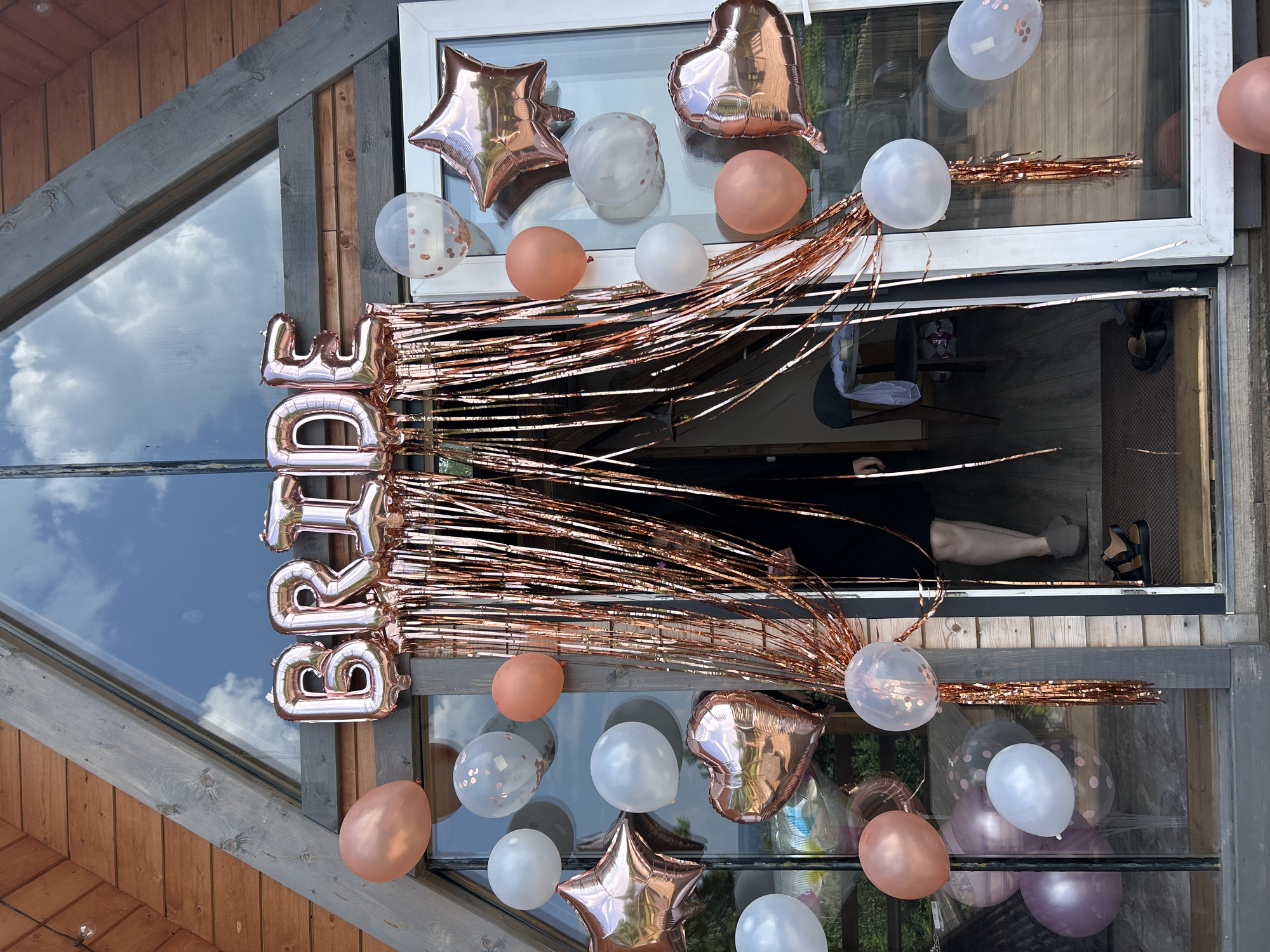 A rustic wooden A-frame building, possibly a house or cabin, is decorated for a bridal event. The main entrance features a large window and a door, both adorned with rose gold balloons and foil decorations. Large foil balloons spelling out "BRIDE" are suspended above a shimmering rose gold fringe curtain that hangs across the entrance. Various other balloons, including round and star-shaped ones, in rose gold, white, and clear with confetti, are attached to the door and window frames. A person, visible from the waist down, stands partially obscured by the fringe curtain, wearing a black dress and dark shoes. The environment appears to be outdoors, with reflections of trees and sky visible in the glass. The specific location in Chaikovsky, Russia, is not discernible from the visual information provided in the image.