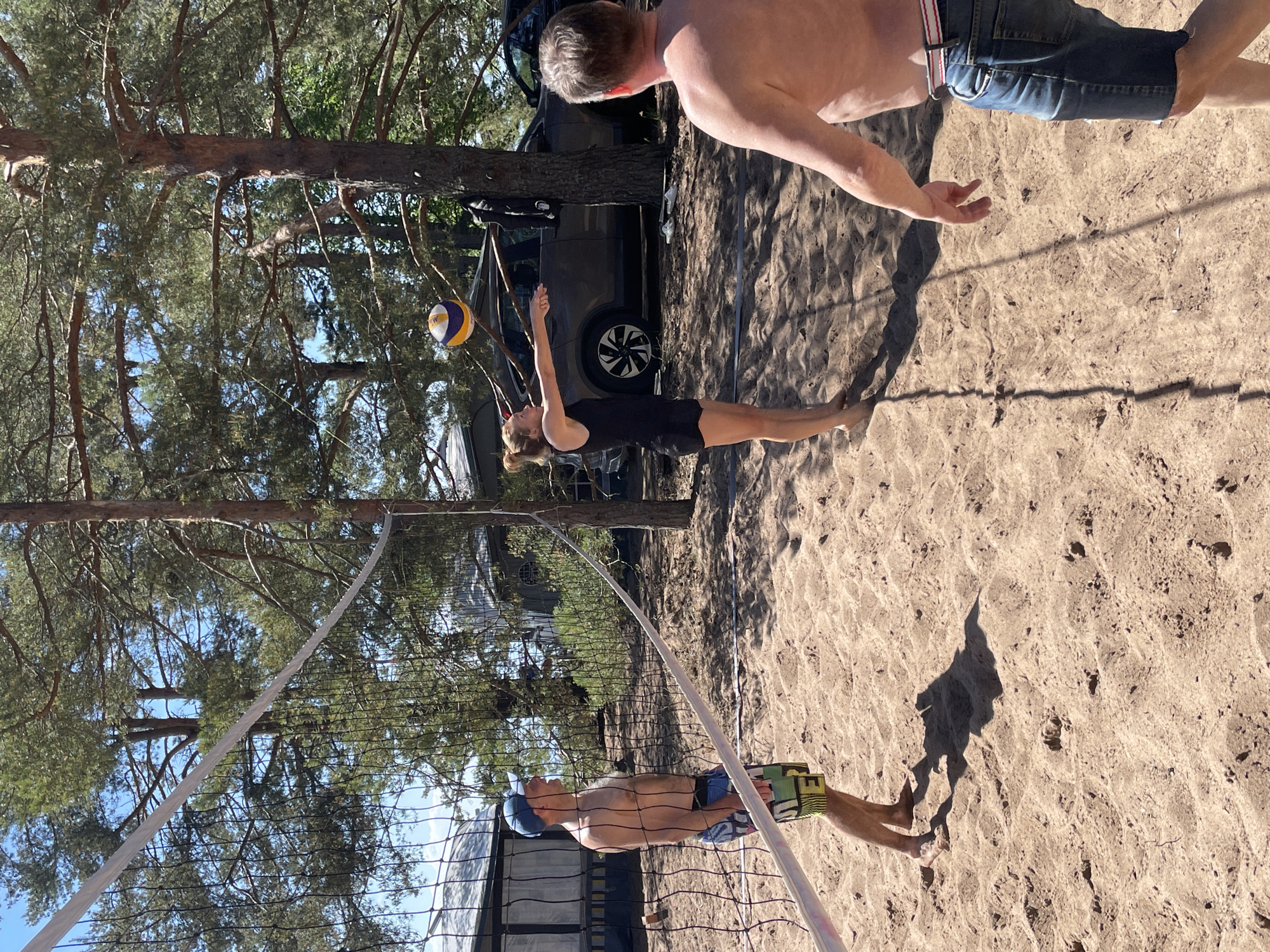 The image depicts an outdoor beach volleyball game in progress on a sandy court in Zelenaya Roshcha, Russia. The scene is illuminated by bright sunlight under a clear blue sky, with numerous mature coniferous trees providing a natural backdrop and partial shade.
Three individuals are present on the court. On the left, a shirtless male wearing a blue baseball cap and multi-colored swim shorts stands barefoot, observing the game. In the center, a female player, dressed in a black tank top and shorts, is captured in the act of performing a forearm pass, with her arms extended and hands joined, focusing on a yellow, blue, and white volleyball suspended in mid-air slightly above her hands. She is also barefoot. On the right, partially viewed from the back, a shirtless male wearing blue denim shorts with a red and white striped belt stands barefoot, observing the play.
In the background, behind the players and trees, a dark-colored SUV is parked, with a dark garment hanging from a tree trunk nearby and a pair of white flip-flops resting on the sand. Portions of residential structures with dark roofs are visible through the foliage. The sandy court exhibits footprints and disturbed areas consistent with active play.