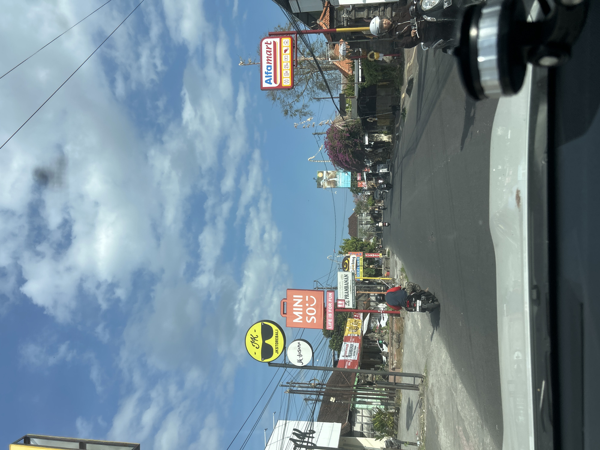 The image depicts a street scene in Sukawati, Indonesia, with various shops and signage visible. On the left side of the road, a building with "JK" signage is prominent, accompanied by a yellow circular sign featuring sunglasses and the text "JKSTOREBALI". Below this, a smaller white circular sign reads "JK.busana". Further down the street, a red sign with white lettering states "MINI SO" and "LIFE IS FOR FUN". To the right, a red and white sign for "Alfamart" is visible. Several motorbikes are on the road, with two riders clearly visible: one wearing a blue and red top, and another wearing a brown jacket and white helmet. The road is paved and appears to be a busy commercial area. The sky is blue with scattered clouds.
