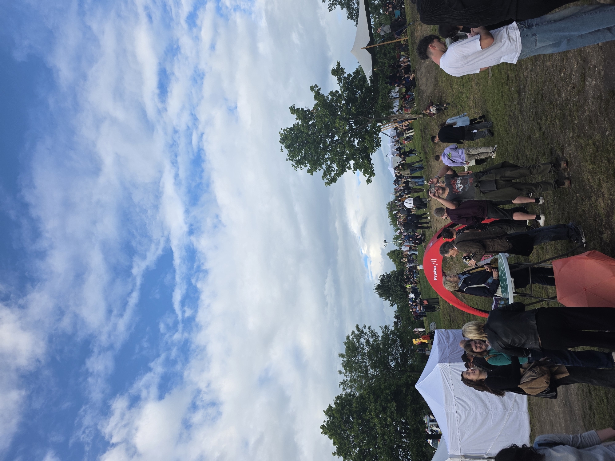 The image depicts an outdoor daytime gathering or festival on a sloping, grassy terrain under a partly cloudy blue sky. **Environment:** The upper two-thirds of the image show a blue sky interspersed with numerous white, cumulus-like clouds. The ground consists of green grass with visible patches of bare earth or dirt paths, indicating foot traffic. Several mature deciduous trees with dense green foliage are prominent, particularly on the left and right sides of the mid-ground. The landscape gently slopes upwards from the foreground towards the background, where a larger crowd is situated on the elevated ground. **Structures and Objects:** - In the left foreground, two white canopy tents, one larger than the other, are erected. - To the right of the white tents, a red arched tent with "Praha ///" branding is visible. A round, white, high-top table with items on its surface stands next to this red tent. - An orange umbrella, folded, lies on the ground in front of the red tent. - On the far right mid-ground, a large, light-colored canvas tent or sunshade is set up near a tree, with people seated underneath it. - In the distant background on the right, what appear to be tall poles with lights are visible. **People:** Approximately 50-70 individuals are visible throughout the scene, primarily adults, dressed in casual attire. - **Foreground (left):** A person with dark hair, seen from the back, wearing a gray top, is partially visible. - **Foreground (center-left, near white tents):** A group of four women are engaged in conversation. One woman, smiling, wears a black long-sleeved top and carries a brown shoulder bag. Another woman, also smiling, wears a green long-sleeved top and dark pants. A third woman wears a black leather jacket and dark pants, facing the group. - **Foreground (center-right, near red tent):** A group of three individuals—two women and one man—are standing around the white table. One woman wears a dark blue jacket and light pants, holding a glass. A man in a brown jacket and dark pants holds a cup. - **Mid-ground (center-right):** A man wearing a grey t-shirt, olive-green cargo pants, and sunglasses is holding a smartphone up to take a photograph or video. Another man in a purple hoodie and beige pants walks behind him. - **Mid-ground (far right):** A man in a white t-shirt and blue jeans is looking down at his smartphone. Beside him, a person