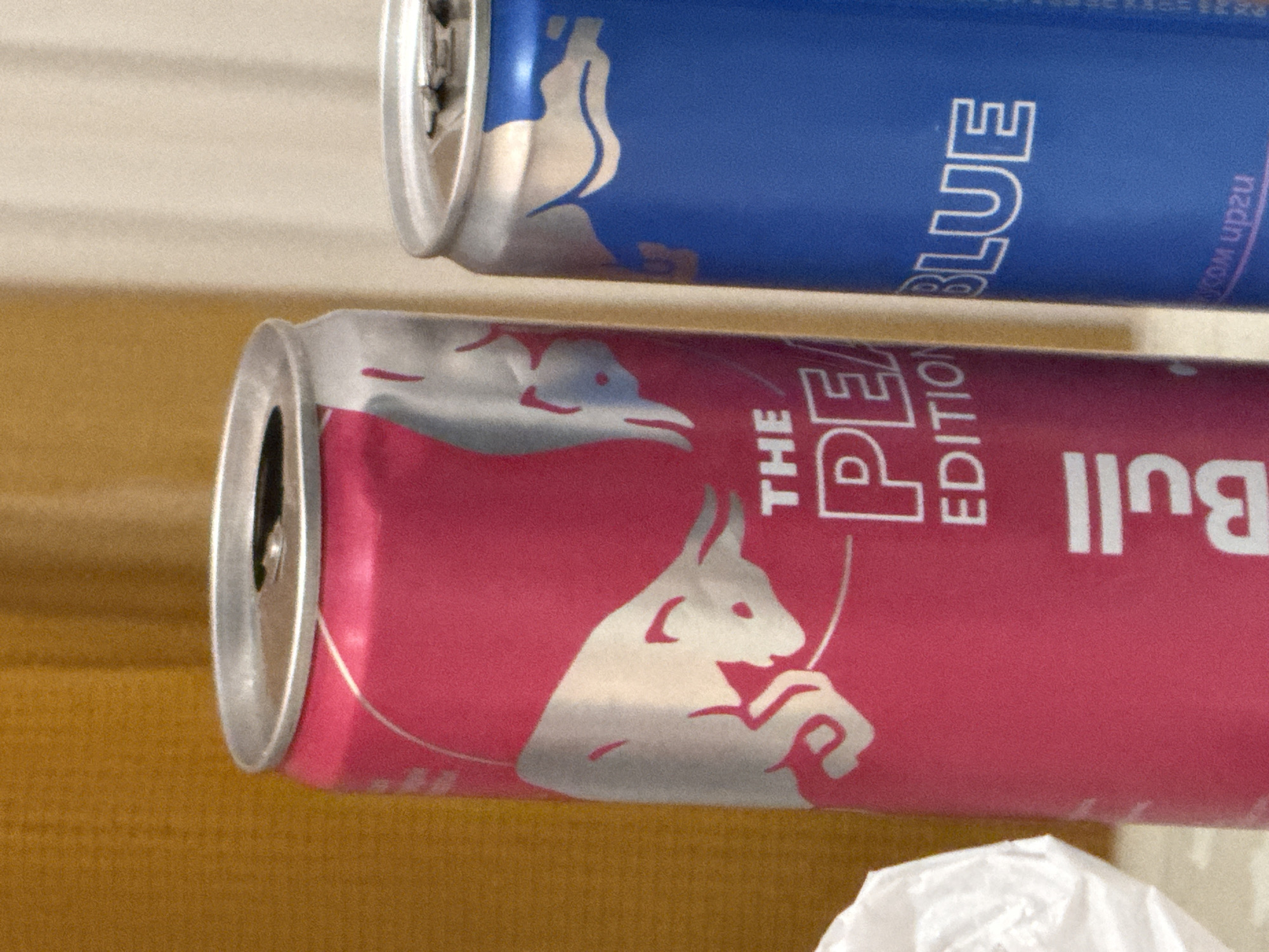 The image shows two Red Bull energy drink cans on a table. The can on the left is pink and has "THE PEACH EDITION" printed on it. The can on the right is blue and has "BLUE EDITION" printed on it. The background is a blurred yellowish-brown wall. There is no indication of the location in the city of ЖК «Борисоглебское», Russia.