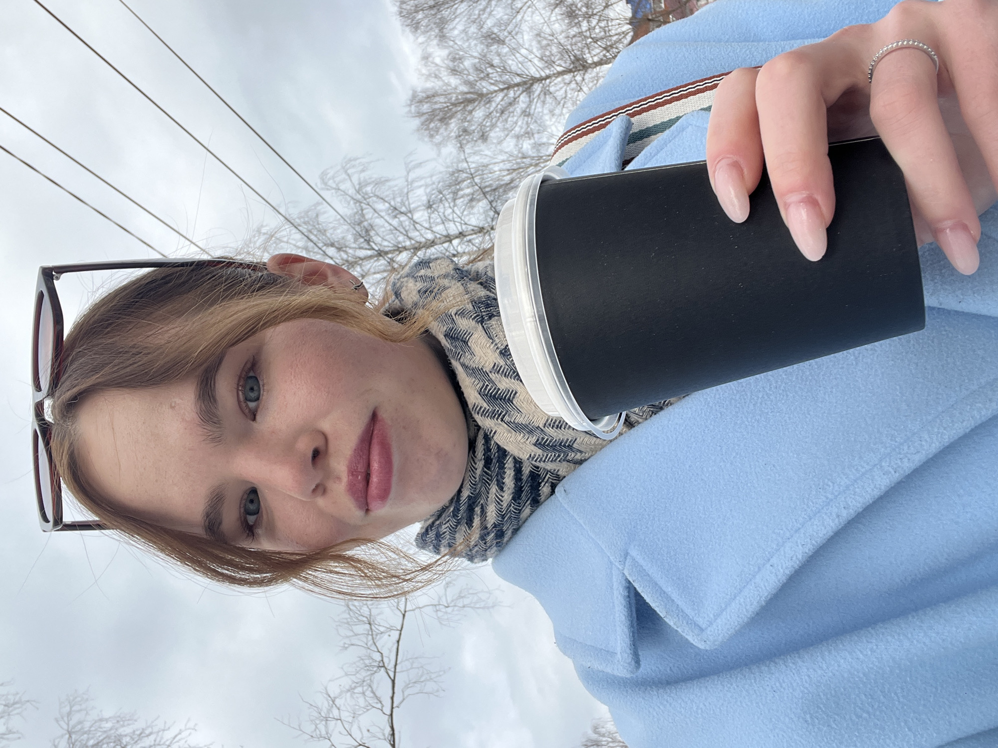 A young woman is holding a black paper cup with a white lid in her right hand. She is wearing a light blue coat and a scarf with a beige and gray pattern. The woman's left hand is visible, and she is wearing a silver ring on her ring finger. She has light skin, light brown hair with blonde highlights, and wears glasses perched on her head. The background consists of a cloudy sky, bare trees, and power lines. The location is Chaikovsky, Russia.