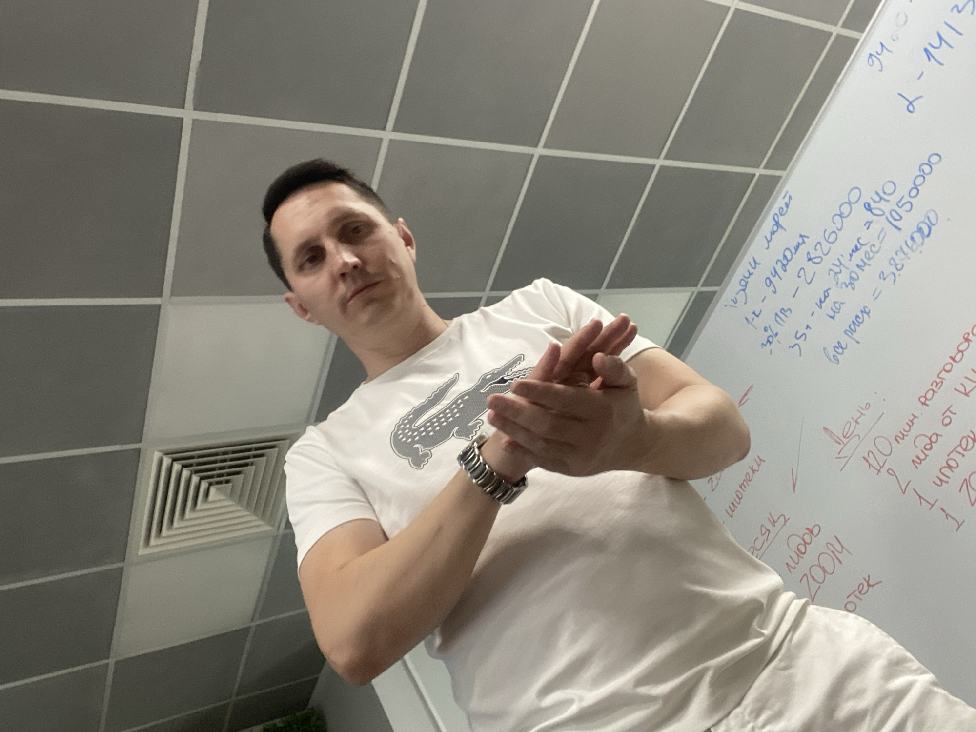 A man in a white t-shirt stands in front of a whiteboard with handwritten notes in Russian. He is looking down at the board. The room has tiled walls and ceiling. The whiteboard contains mathematical calculations and lists, possibly related to business or personal finance. The man is wearing a watch on his left wrist. The scene appears to be indoors, possibly an office or meeting room.