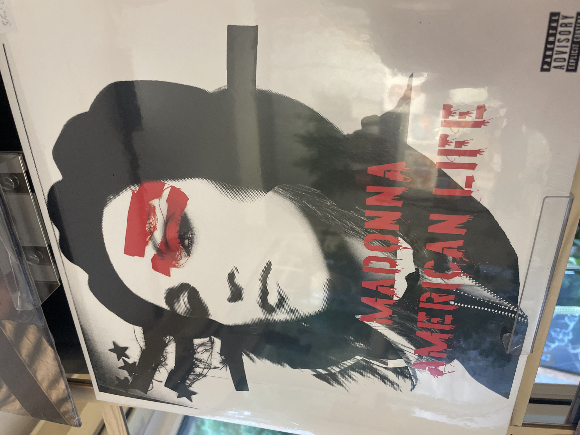 The image shows a vinyl record album cover for Madonna's "American Life". The cover features Madonna's face with red markings across her eyes. The text "MADONNA AMERICAN LIFE" is printed in red vertically on the right side. A "Parental Advisory Explicit Content" label is visible in the top right corner. The album is displayed in what appears to be a record store or collection, likely for sale or display. The image does not provide any information about the location being in Novi Sad, Serbia.