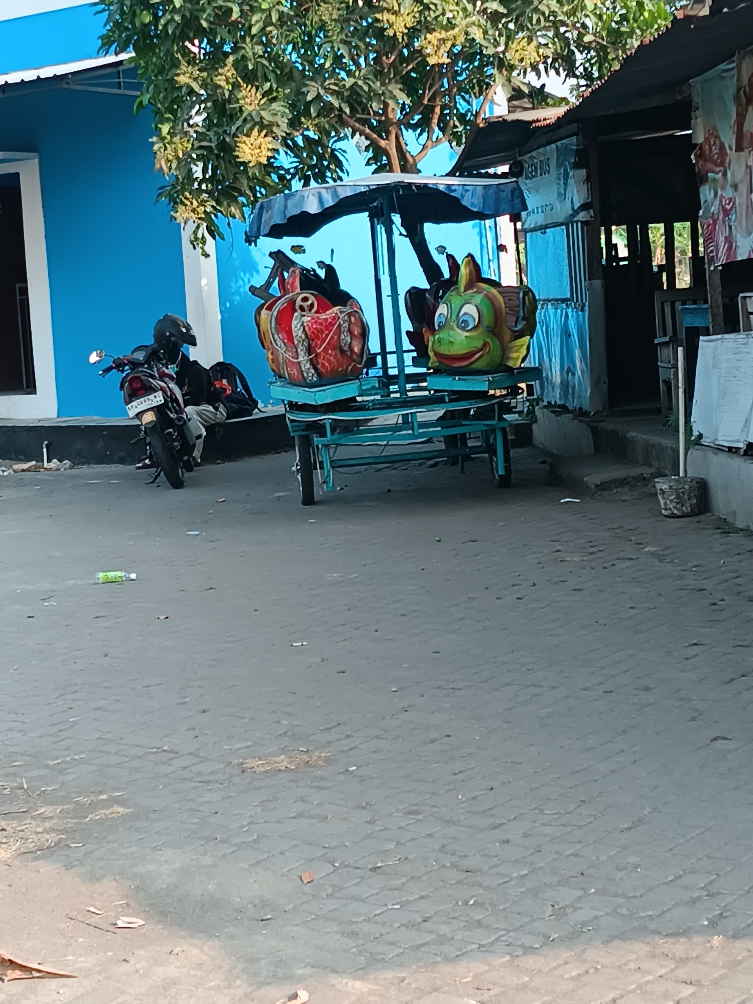 In Imogiri, Indonesia, a blue building stands on the left, partially obscured by a leafy tree. In front of the building, a motorcycle is parked. To the right of the motorcycle, a colorful cart with two cartoonish figures, one resembling a frog and the other a plump red creature, is positioned. The cart has a blue canopy and is situated on a paved area. Behind the cart, a small structure with a corrugated metal roof is visible. The scene appears to be outdoors, possibly in a public area.