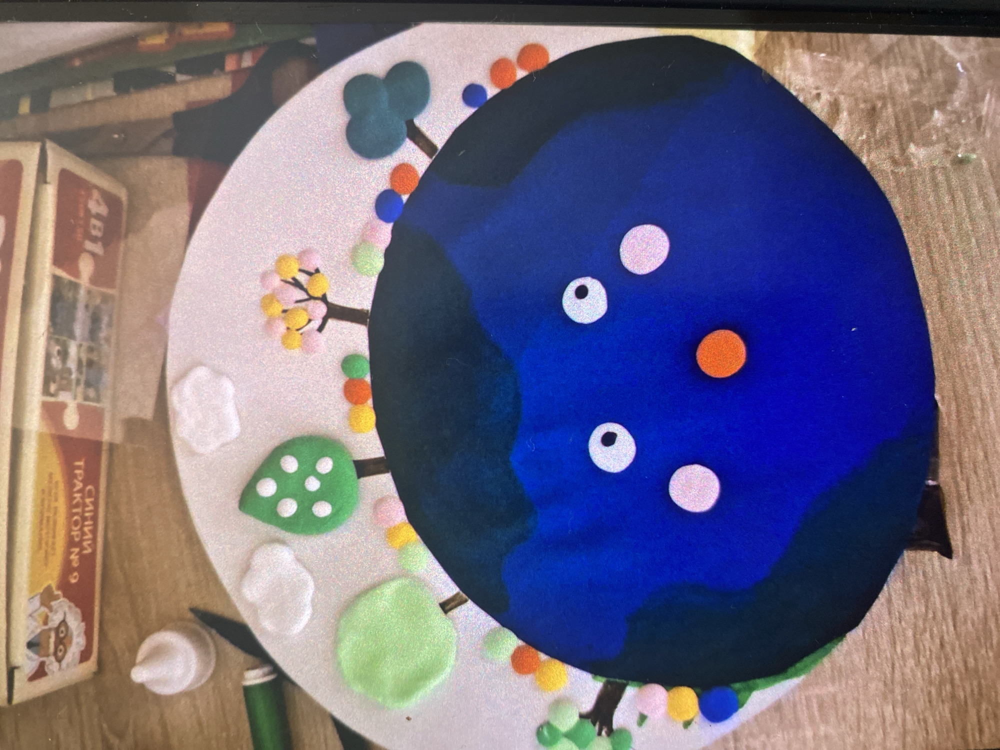 The image displays a whimsical, circular decorative object, possibly a cake or a craft project, set on a white base. The central feature is a large, dark blue circle, resembling a stylized planet or cartoon face, with lighter blue patches suggesting landmasses or shading. It features three white circular "eyes" (two with black pupils) and one orange circular "mouth."
Around the perimeter of the white base, various colorful decorations are arranged. These include small, fluffy pom-poms in shades of pink, yellow, orange, blue, and green, attached to brown stick-like elements, creating an impression of miniature, blossoming trees. Interspersed among these are flat, green leaf-like shapes, some with white polka dots, and white cloud-like cutouts, adding to a playful, natural scene.
In the background, a wooden surface is visible, likely a table or floor. To the left, a cardboard box with Cyrillic text is partially seen. The visible text reads "ТРАКТОР № 9" (TRAKTOR No. 9) and "СИНИЙ" (SINY, meaning BLUE), along with the number "481" on the top flap. A cartoon image of a white-bearded figure is also visible on the box. Near the bottom left, a small white cap and a green object, possibly a tube or craft tool, rest on the wooden surface.
The overall impression is one of a creative project or celebration item, possibly for a child, characterized by bright colors and simple, charming shapes. Given the Cyrillic text and location context, the scene is set in Saint Petersburg, Russia.