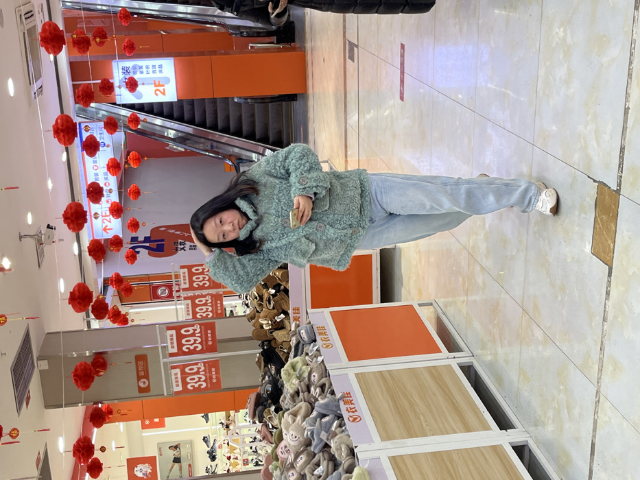 An indoor, well-lit retail environment in Jiangjin, China, displays numerous red Chinese lanterns suspended from the ceiling, indicating a festive period. The tiled floor features a light, possibly off-white pattern with occasional darker square inserts. In the center, a female of East Asian descent stands, facing slightly left with her gaze directed leftward. She wears a teal, textured, fuzzy jacket with visible buttons and light-wash blue jeans, paired with white sneakers. Her right hand is raised, touching her long, dark hair, while her left hand holds a light-colored smartphone. Her mouth is slightly ajar with a neutral to contemplative expression.
To her immediate left, two white and orange display bins are filled with various soft slippers, some featuring animal designs. Red signs above these bins display "39.9" (likely a price in Yuan) and Chinese characters, including "超值单品" (great value item). Behind these bins, more footwear is visible on lower shelves. On the far right, a dark-colored escalator ascends to an upper floor. Adjacent to the escalator, an orange pillar displays a white sign with "2F" (second floor) and an upward arrow, alongside Chinese characters listing departments such as "女装" (women's clothing), "针织" (knitwear), and "内衣" (underwear). A partial figure of another person, clad in dark attire and holding a smartphone, is visible on the far right edge of the frame.