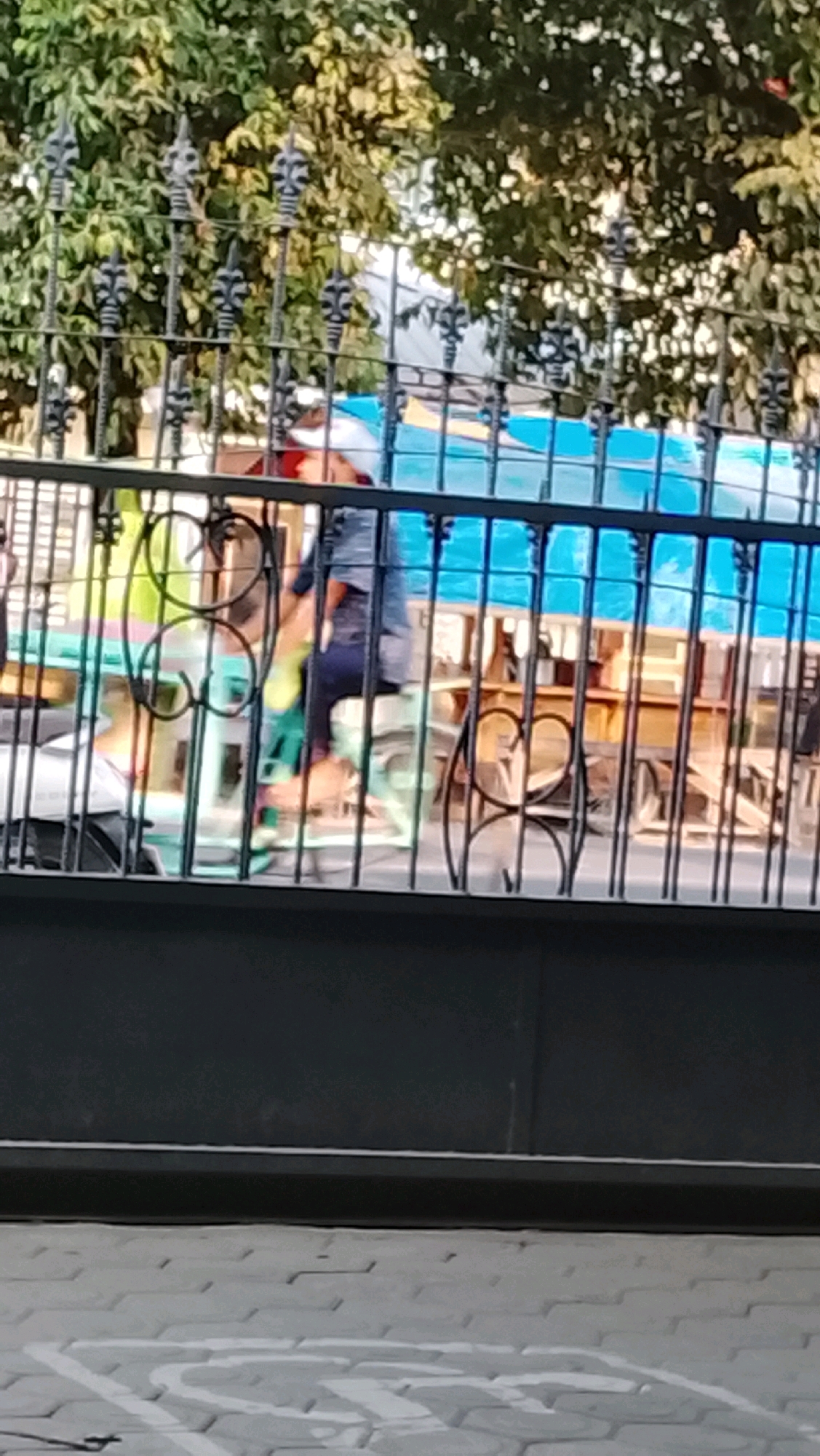 The image shows a person on a bicycle, viewed through a black metal fence. The person is wearing a blue shirt and dark pants, and is riding a bicycle with a green frame. The background consists of trees and a blue awning. The scene appears to be outdoors, possibly in a residential area. It is not possible to determine the exact location within Slawi, Indonesia, from this image alone.