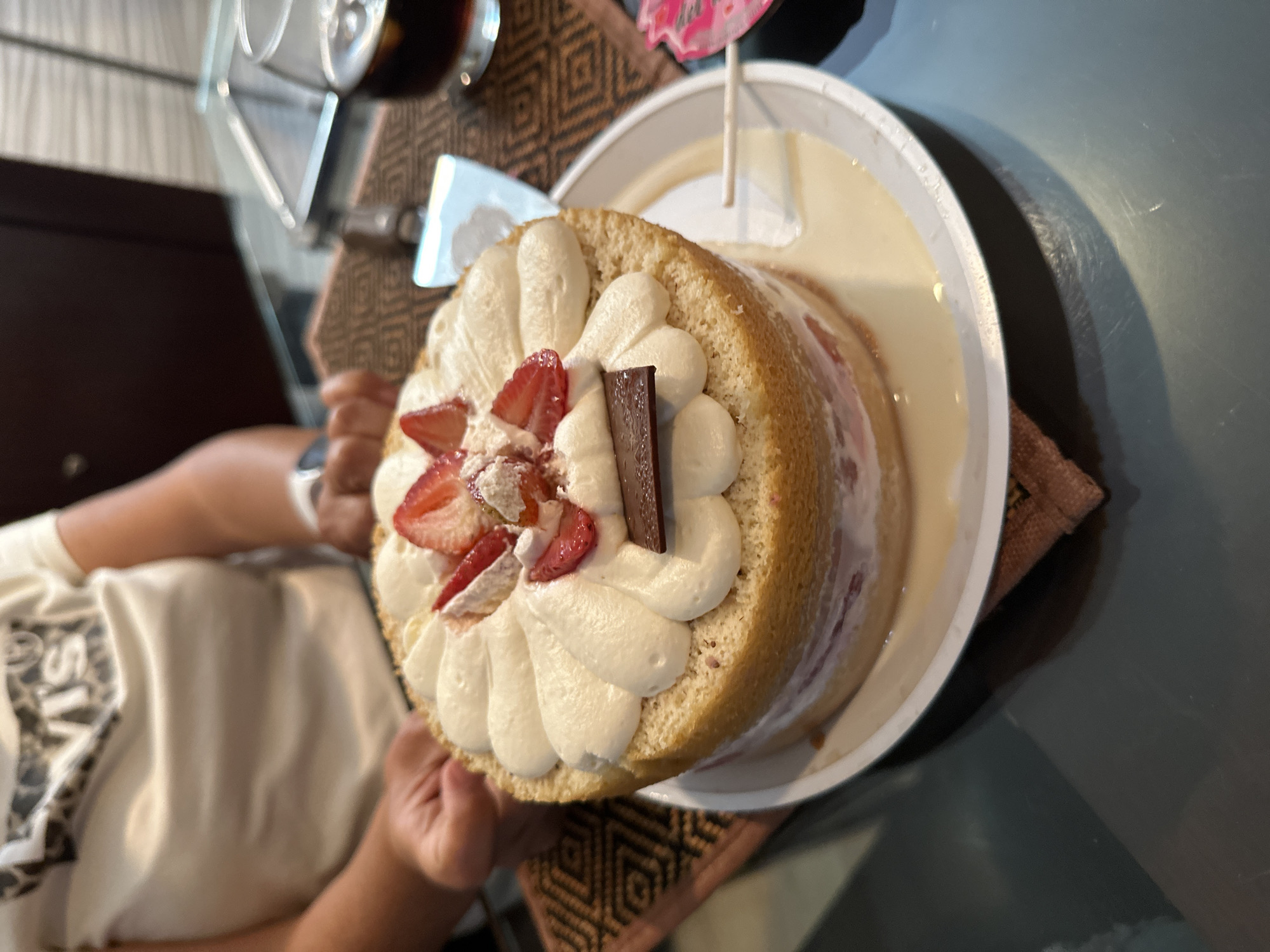 A medium-high angle shot shows a person holding a white plate with a cake in it. The cake is a round sponge cake covered with white frosting rosettes, topped with slices of strawberries and a chocolate stick. The cake is sitting in a pool of pale yellow liquid, likely tres leches sauce. A small pink cake topper is visible. The person holding the cake is wearing a light-colored t-shirt with writing on it. Their arm and hand are visible, holding the plate. The background shows a glass table with a woven placemat underneath, and a glass with a dark liquid inside. The scene appears to be indoors, possibly a dining area. Information about the specific location in Puebla, Mexico is not present in the image.