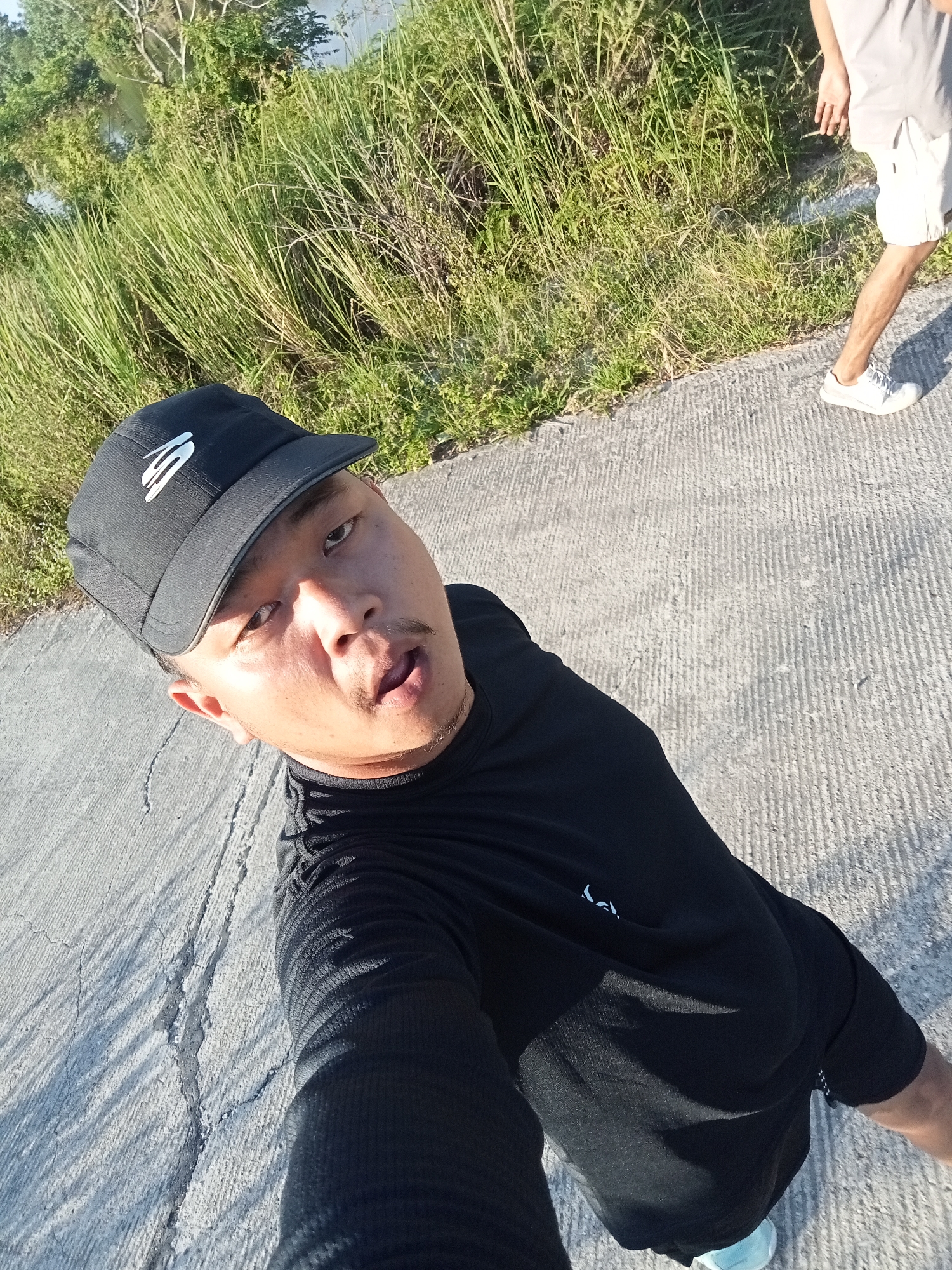 A young Asian male wearing a black baseball cap, black t-shirt, and black shorts is captured from a high angle, looking up at the camera. He is standing on a paved surface next to a grassy area with trees and a body of water visible in the background, suggesting an outdoor location, possibly along the river in Kualu, Indonesia. Another person, wearing white shorts and white sneakers, is partially visible on the right side of the frame, also on the paved surface. The lighting indicates it is daytime, with shadows cast on the ground.