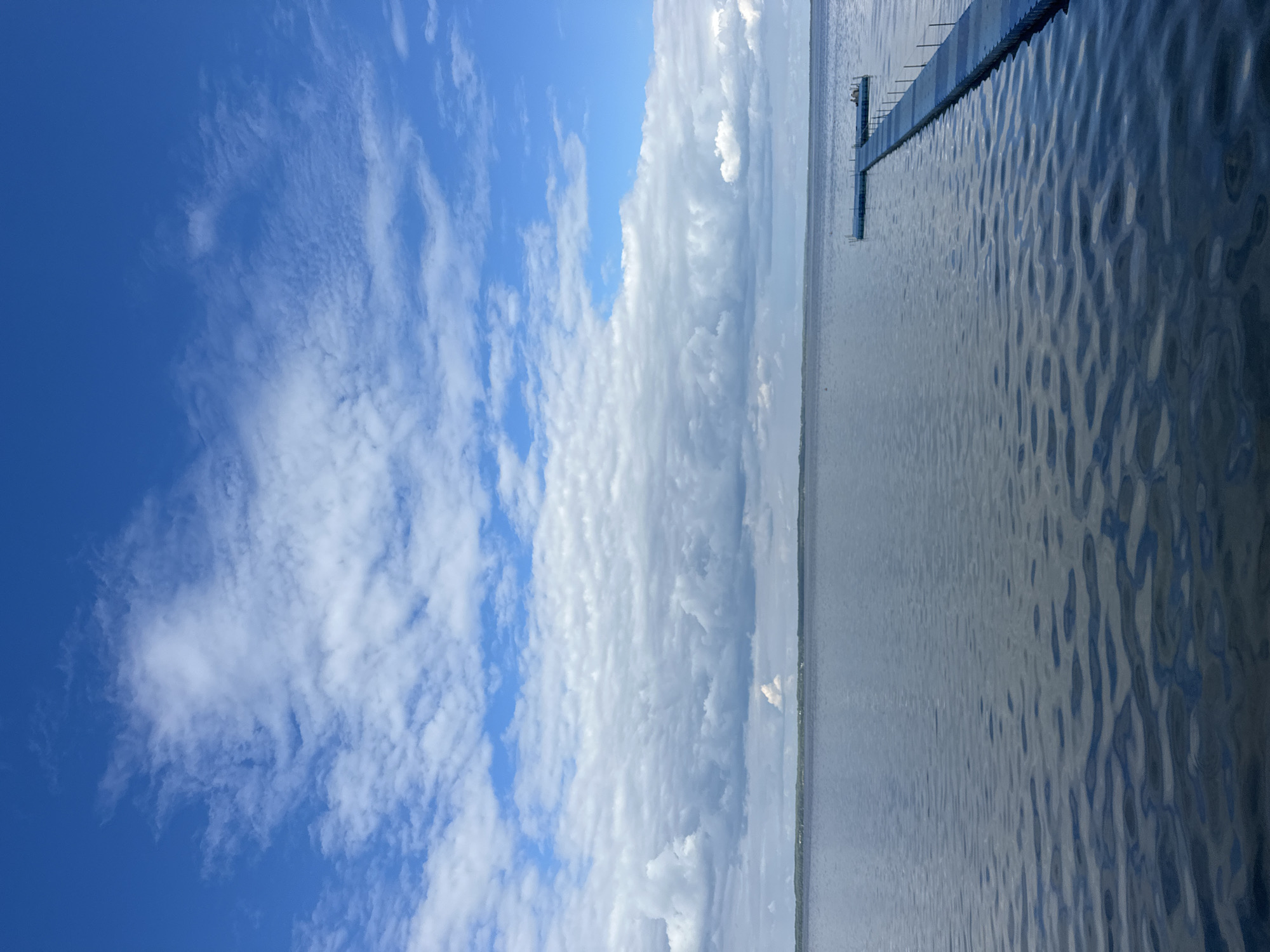 The image depicts a serene lake under a vast, partly cloudy blue sky. In the foreground, the calm water shows gentle ripples. A blue modular dock extends from the right side of the frame, jutting out into the water. At the end of the dock, there appears to be a person or two sitting. The horizon line is low, with a distant tree line and structures visible on the far shore, suggesting a rural or natural setting. The overall atmosphere is peaceful and expansive. This scene is located in Narachansky Selsoviet, Belarus.