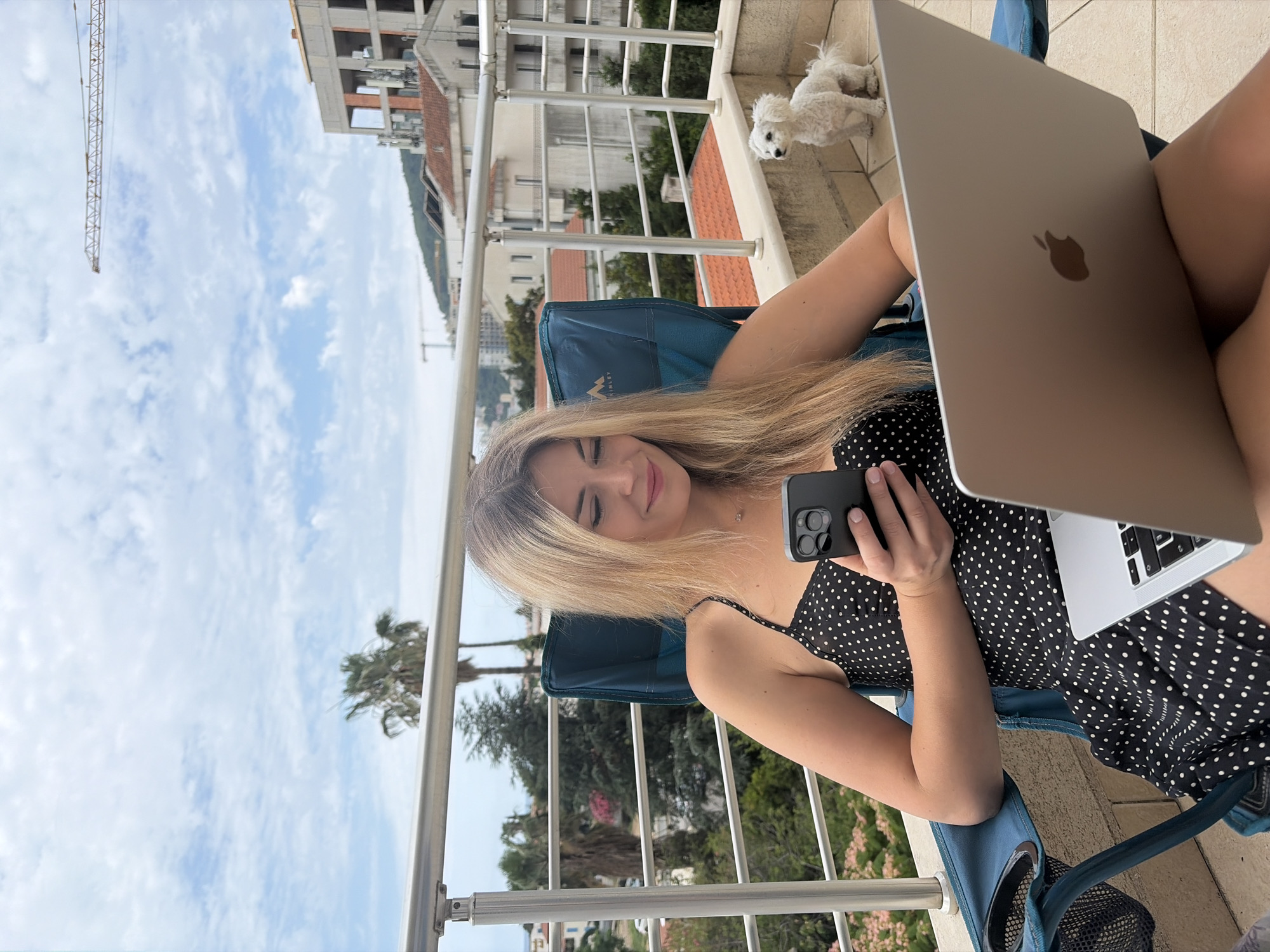 A young woman with long blonde hair is depicted relaxing on a balcony, likely in Budva, Montenegro. She is seated comfortably in a blue folding chair, wearing a black polka-dot dress. On her lap rests an open silver MacBook laptop, displaying the Apple logo on its lid. In her left hand, she holds a dark-colored smartphone with a visible triple-camera array, at which she gazes with a gentle smile and partially closed eyes, conveying a relaxed and content mood.
In the upper right background, a small, white, fluffy dog stands on the tiled balcony floor, observing the scene. The balcony features a metal railing that frames a panoramic view of a town. The landscape beyond includes numerous buildings with distinctive red-tiled roofs, interspersed with lush green trees and several palm trees. Distant hills or mountains are visible under a bright, partly cloudy sky. A prominent construction crane extends upwards in the background, suggesting ongoing development.
The brand "MCKINLEY" is visible on the blue fabric of the woman's chair. The overall scene suggests a pleasant daytime setting, where the woman is enjoying a moment of leisure or remote work in a scenic, Mediterranean environment.