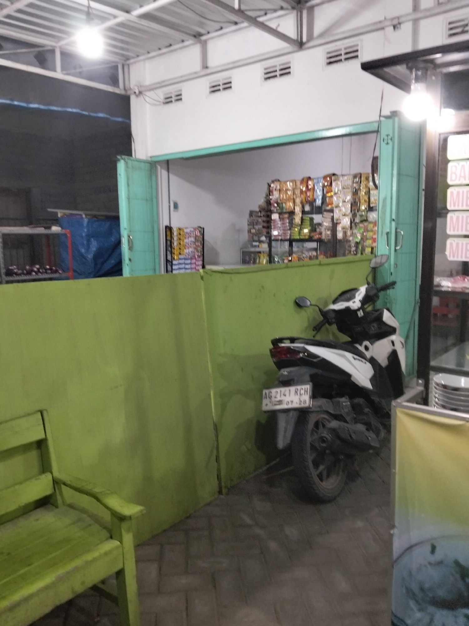 The image depicts an outdoor scene in Gempolan, Indonesia. In the foreground, a bright green wooden bench is partially visible on the left. Behind the bench, a light green wall dominates the middle ground. To the right of the wall, a white and black motorcycle is parked. The motorcycle has a license plate that reads "AG 2141 RCH 07-28". Further to the right, a structure with a glass front displays stacks of plates and a yellow banner. In the background, shelves stocked with various packaged goods are visible behind a light green door. The scene appears to be an open-air establishment, possibly a shop or food stall, with a covered roof structure overhead.