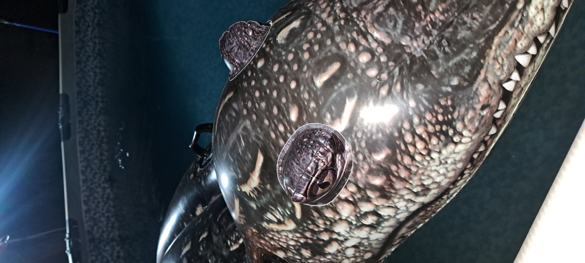 An inflatable alligator toy floats in dark water, possibly a pool or pond. The toy's textured, scaled surface is detailed with dark brown and cream-colored spots. Two round, cartoonish eyes are attached to the top of its head, one on the left and one on the right, with visible pupils. A black plastic handle is secured to the top of the toy, near the center. The background is dark blue, and a bright light source is visible in the upper left corner, casting a glare on the toy. The scene does not provide any information about the location in the city of Азьмушкино, Russia.