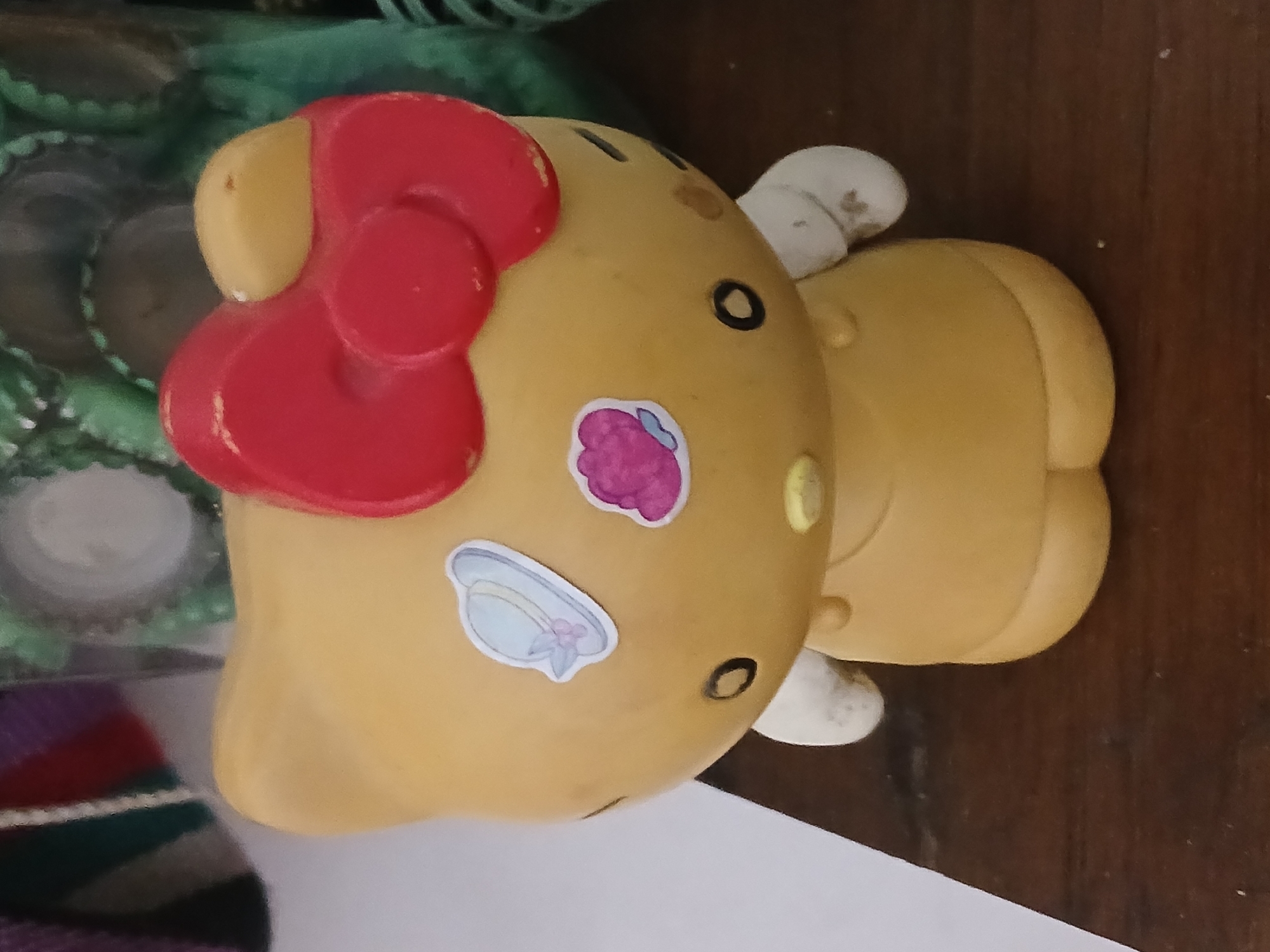 The image captures a yellow, Hello Kitty-shaped object lying on its side, likely a piggy bank or decorative item, in Buenos Aires, Argentina. The object features a prominent, glossy red bow on its head, with characteristic black oval eyes and a small yellow oval nose. Two white, ear-like protrusions are visible on its head, and two small stickers adorn its body: one depicting a pink, berry-like shape and another showing a light blue hat with a pink bow.
The background consists of a dark wooden surface to the right and beneath the object, suggesting it rests on a table or shelf. To the left, a blurry green plastic or mesh material is visible, along with several metallic bottle caps, indicating a somewhat cluttered environment. Part of a striped fabric, with visible hues of purple, red, and dark green, is partially seen in the lower-left corner. The lighting suggests an indoor setting. No activity is occurring, and there is no discernible text on the object or in the surroundings.