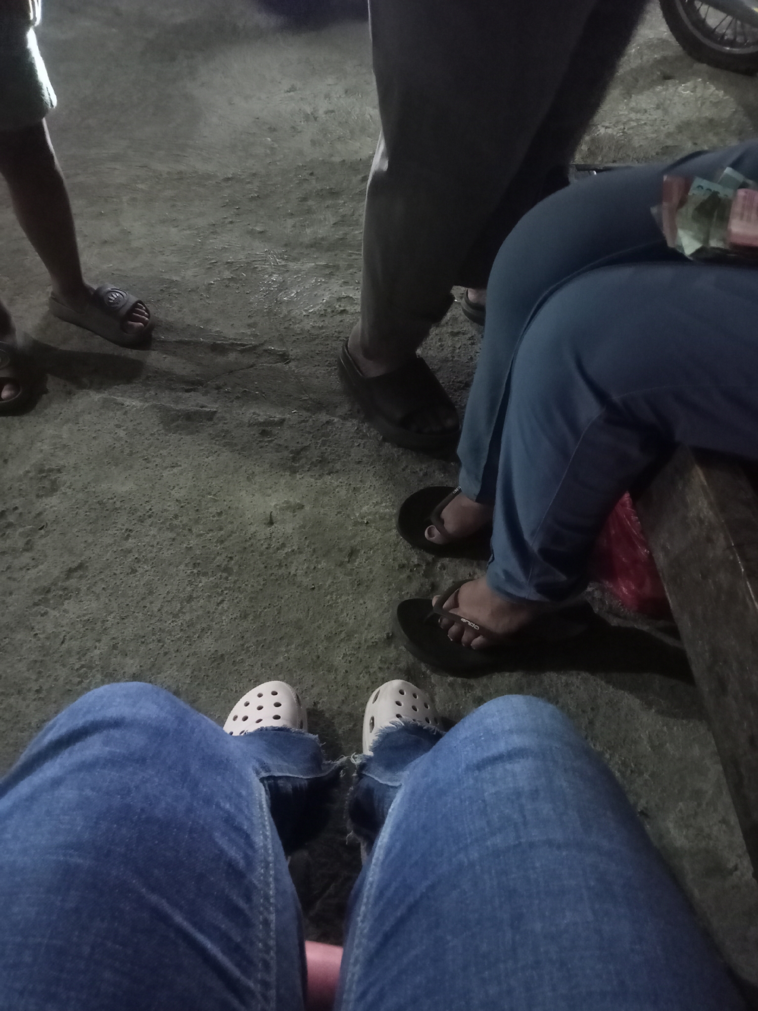 The image shows a ground-level view of four individuals' legs and feet, positioned in varying orientations. The ground appears to be a rough, textured concrete surface, possibly outdoors.

In the foreground, a person wearing blue jeans and white Crocs is positioned with their legs extended forward and slightly apart. To the left and slightly behind, another individual's legs are visible, also wearing blue jeans. Their feet are clad in white Crocs with distinct circular perforations.

Further back and to the right, a person is seated, their legs covered in blue fabric, likely pants. They are wearing dark flip-flops. A glimpse of their hand, holding a wad of Indonesian Rupiah banknotes, is visible. To the far right, a wooden bench is partially visible.

In the upper left of the frame, a person's lower leg and foot are visible, wearing a light-colored skirt or shorts and a dark sandal. The scene suggests a casual gathering or waiting area, possibly in a public space. The lighting is dim, indicating it may be evening or nighttime. The location is specified as Tengaran, Indonesia.
