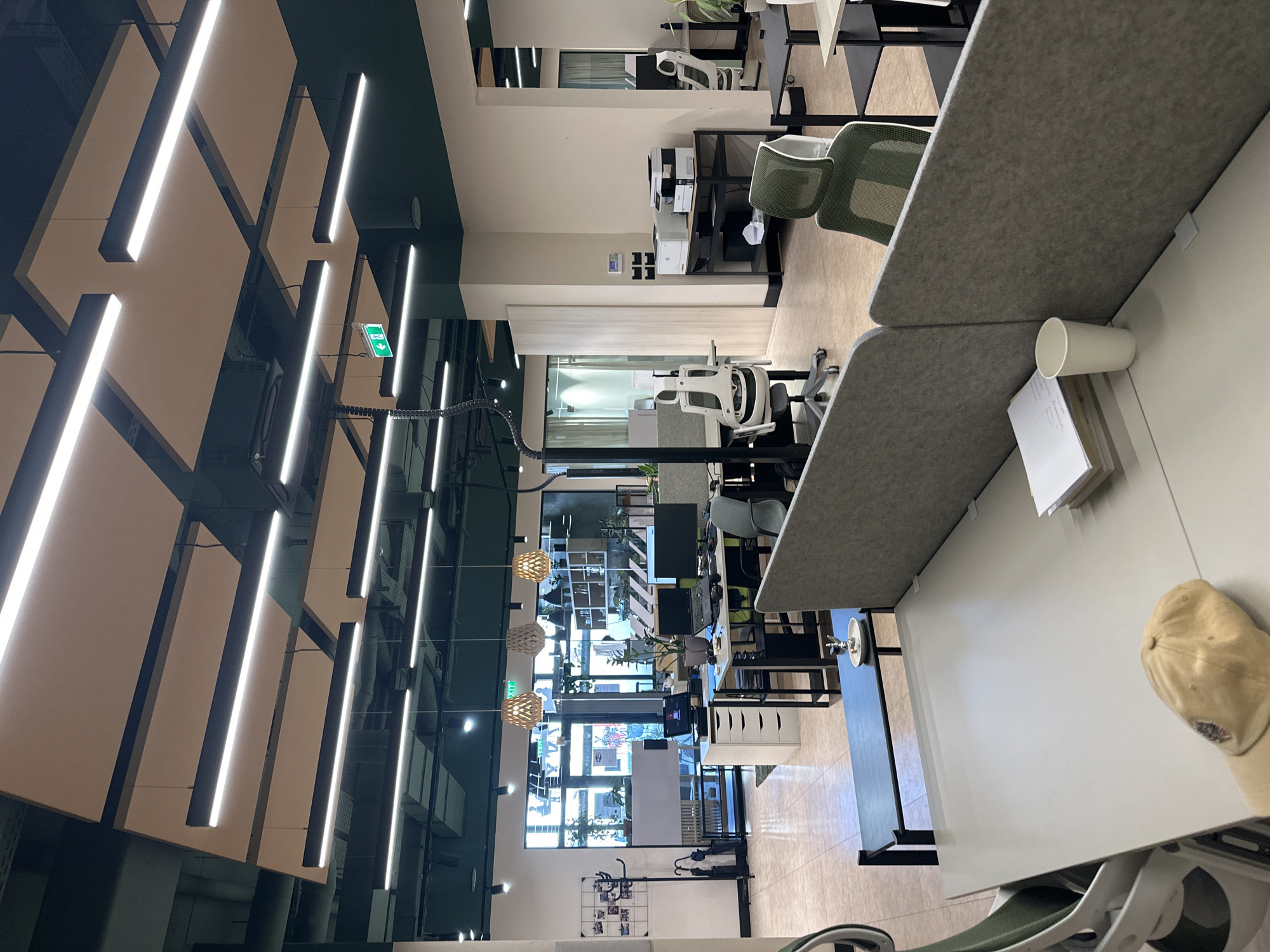 This is a forensic, matter-of-fact description of an office space. The image depicts an open-plan office with multiple workstations. The ceiling features a combination of dark green panels and rectangular acoustic panels with integrated linear LED lighting. The walls are painted in neutral tones, with some areas of dark green trim.
In the foreground, there are two desk partitions made of gray felt material. On the desk to the right, a stack of papers and a white paper cup are visible. A beige baseball cap rests on the desk in the lower-left corner.
Further into the office, various workstations are set up with computers, monitors, and office chairs. Some chairs are white and ergonomic, while others are green with mesh backs. A printer and scanner are situated on a black metal shelving unit. Large windows at the back of the office allow natural light to enter and provide a view of the exterior, including greenery and a glimpse of a street scene. There are also decorative pendant lights with wooden shades hanging from the ceiling. An emergency exit sign with a green arrow is illuminated and visible. The floor is tiled with a light-colored tile. The overall impression is a modern and functional office environment.
The image is captioned as being from Budva, Montenegro.