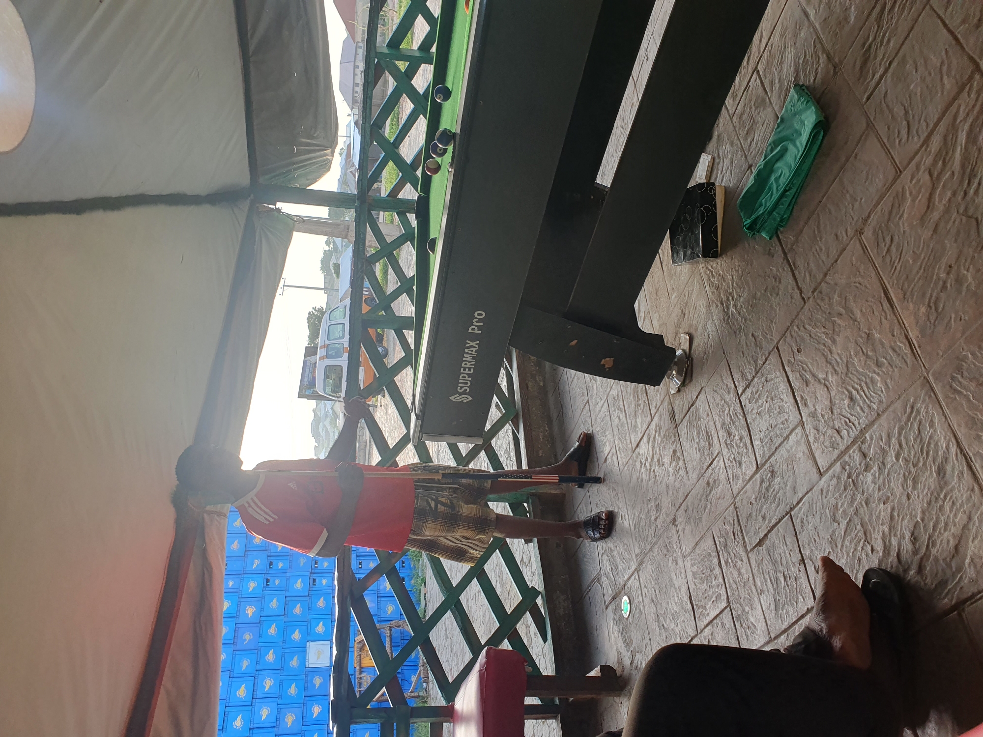 In Lokoja, Nigeria, an outdoor covered patio features a billiard table with several balls on its green felt surface. To the left of the table, a young man wearing a red shirt and plaid shorts stands holding a pool cue. His feet are clad in black sandals. The patio floor is paved with textured rectangular tiles. In the background, stacked blue crates are visible behind a green wooden railing, and a white van is parked further in the distance. The scene is illuminated by natural light, suggesting it is daytime.