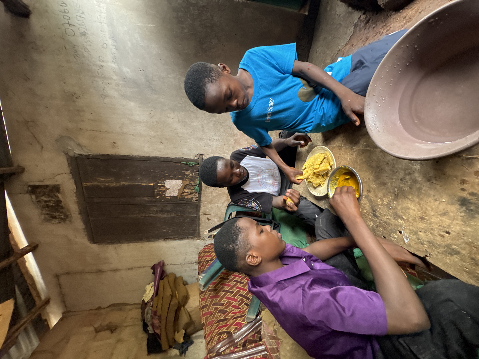 Three boys are eating a meal together in a poorly lit, rustic indoor space. The boy on the left wears a purple shirt and is partially lying down. The boy in the middle wears a white and black shirt. The boy on the right wears a blue shirt. They are sharing plates of food, one plate with a yellow substance and another with a brownish, lumpy substance. A large brown bowl is nearby. The walls of the room appear to be made of rough concrete. There is a wooden door or paneling on one wall. In the background, there is a pile of blankets or clothes. The scene is taking place in Oboama, Nigeria.