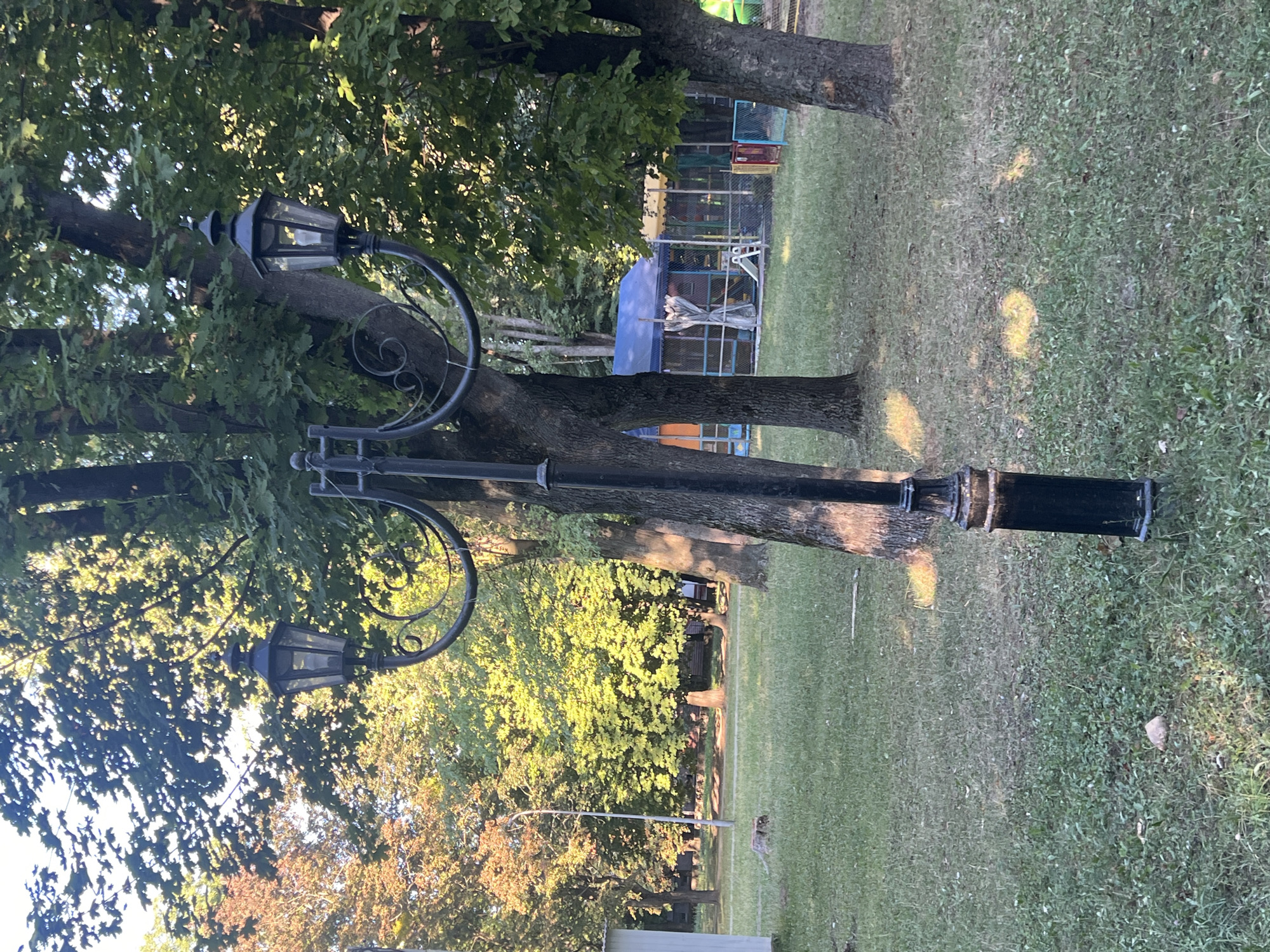 A park scene at ground level shows a black metal lamppost with two lanterns in the foreground. The lamppost is ornate, with curved arms supporting the lanterns. The lanterns themselves appear to be of a traditional design with glass panes. Behind the lamppost, large trees with green foliage dominate the background. Sunlight filters through the leaves, casting dappled shadows on the grassy ground. To the right, a portion of a children's play area with a blue awning and fences is visible. A white building is partially seen on the left side of the image. The location is identified as Sheptytskyi, Ukraine.