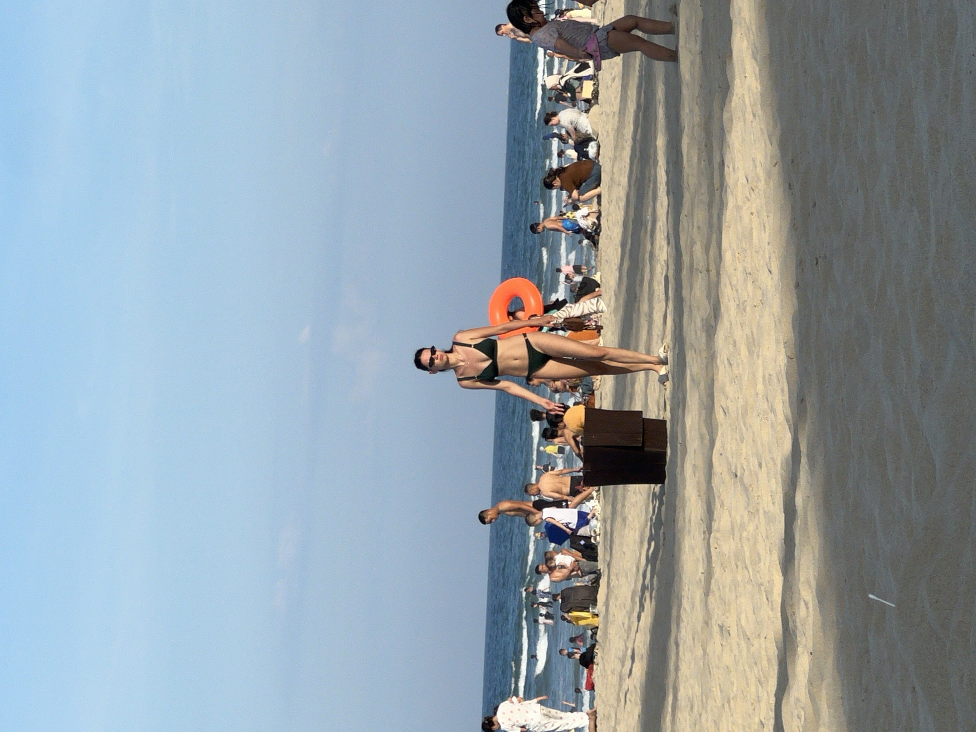 The image depicts a crowded beach scene in Da Nang, Vietnam. The foreground is dominated by sand, with shadows cast by unseen objects. A woman in a dark green bikini walks towards the viewer in the center of the frame. To her left, a large orange inner tube is visible. In the background, the ocean stretches to the horizon, with waves breaking on the shore. Numerous people are scattered across the beach, some sitting, some standing, and some wading in the water. The sky is a clear, light blue.