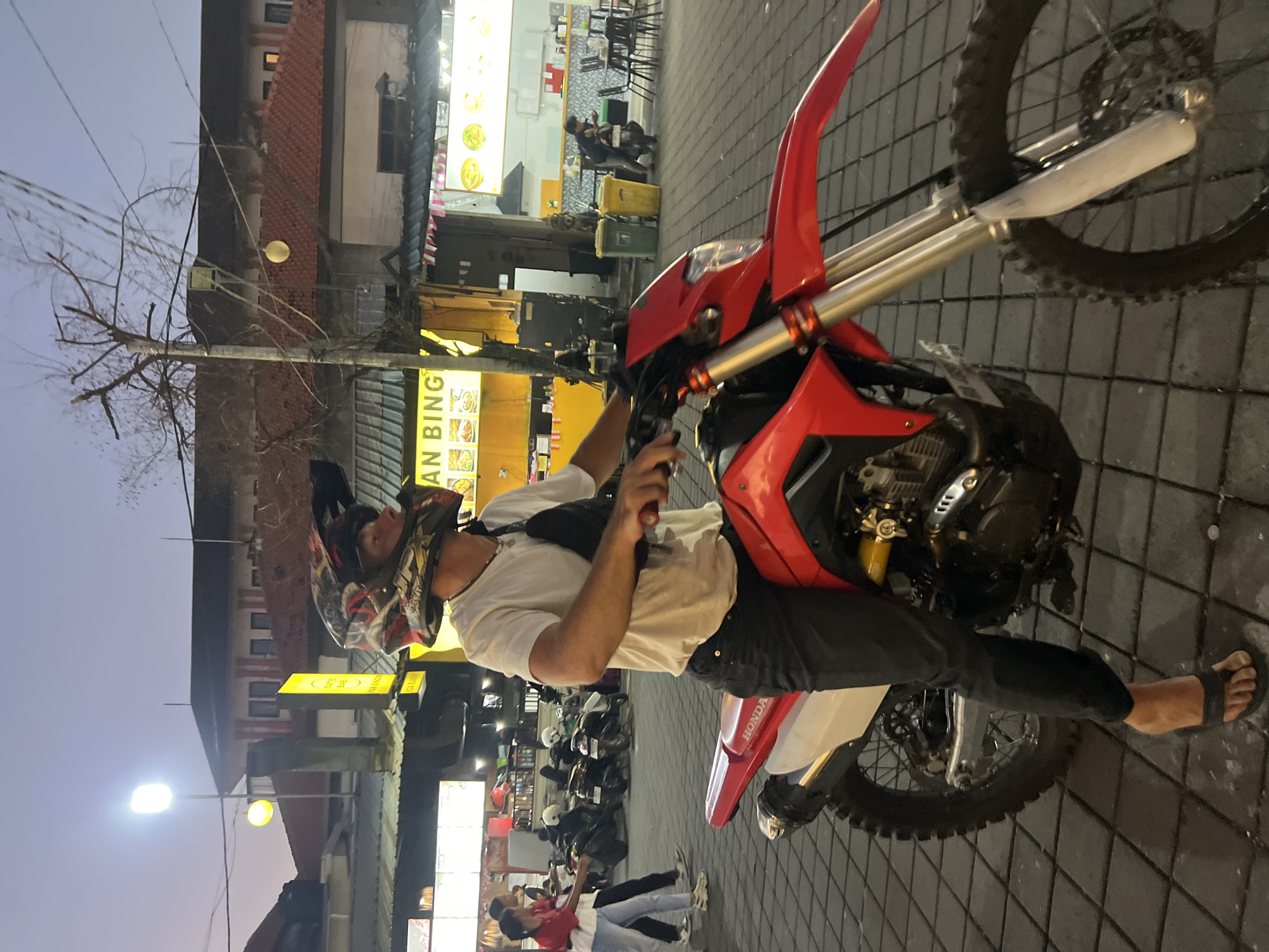 The image depicts a street scene in Kuta, Indonesia, at dusk. A person wearing a red, black, and white helmet, a white t-shirt, black pants, and black flip-flops is seated on a red and white dirt bike. They are looking to their right, with their right hand on the handlebars. A black waist bag is worn across their chest.
In the background, there are several food stalls with yellow signage. One sign reads "AN BING." Buildings with red-tiled roofs are visible. Various motorcycles are parked in the area. To the left of the frame, two individuals, one in a red shirt and white top, and the other in a white top and blue jeans, are walking. Two yellow and green trash bins are situated near a lamp post. The ground is paved with square tiles. The overall lighting suggests it is late evening, with streetlights illuminating the scene.