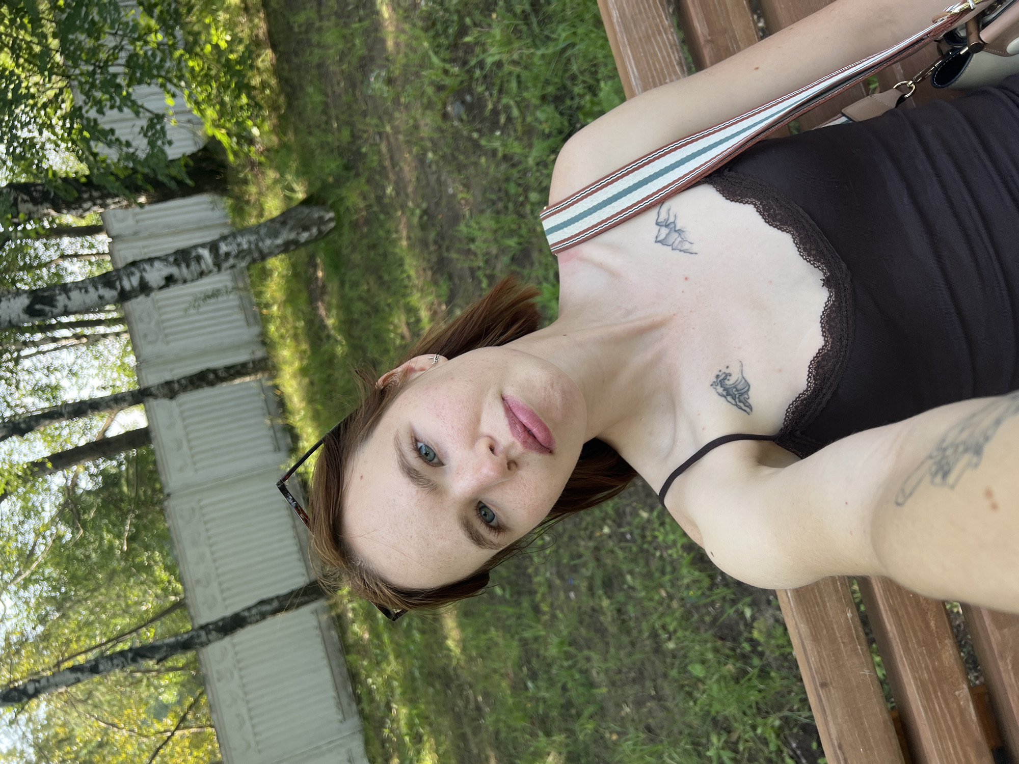 In a park setting in Chaikovsky, Russia, a young woman with short auburn hair and light blue eyes takes a selfie while sitting on a wooden bench. She is wearing a dark brown tank top with lace trim, and a patterned strap from a purse crosses her shoulder. She has several tattoos visible on her chest and arm, including a mountain range and a wave. Her eyeglasses are perched on top of her head. The background consists of trees, green grass, and a white decorative fence. The sun is shining, creating dappled light and shadows on the ground.