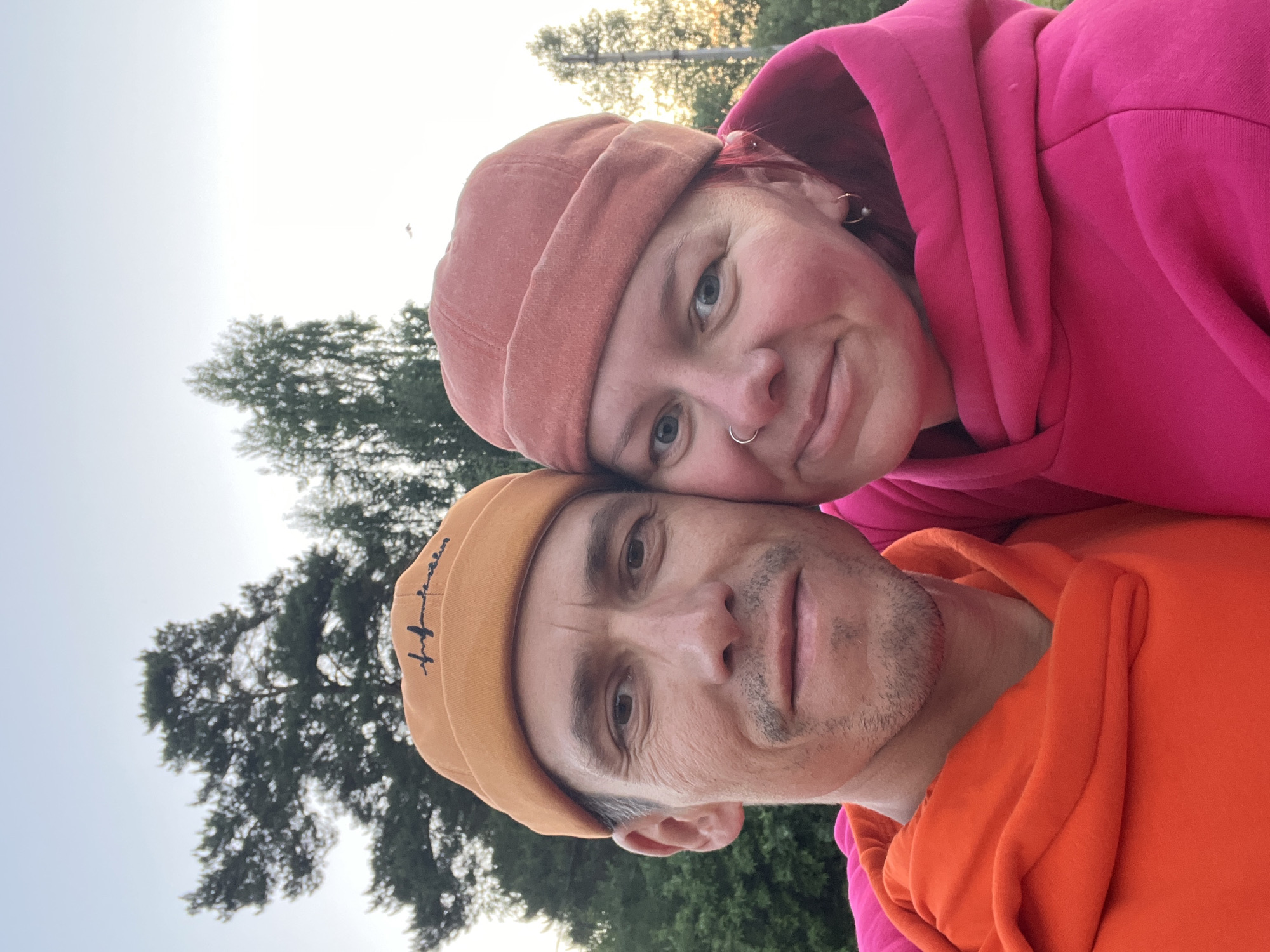 This image captures a close-up selfie of two individuals, a man and a woman, in an outdoor setting in Chaikovsky, Russia. The image is tilted counter-clockwise.
The woman, on the right and slightly higher, has red or pink hair and wears a salmon-colored beanie and a vibrant hot pink hooded sweatshirt. She has a nose ring and hoop earrings, and looks directly at the camera with a gentle smile.
The man, on the left and slightly lower, has light stubble and wears an orange beanie with stylized dark text visible on its front, and a matching orange hooded sweatshirt. His head is leaned against hers, and he also looks towards the camera.
The background features dark green pine trees under a bright, diffused sky, suggesting daytime, possibly late afternoon or early evening due to the soft lighting. Both individuals appear content and affectionate. The text on the man's beanie is a flowing script, resembling a word like "inspiration."