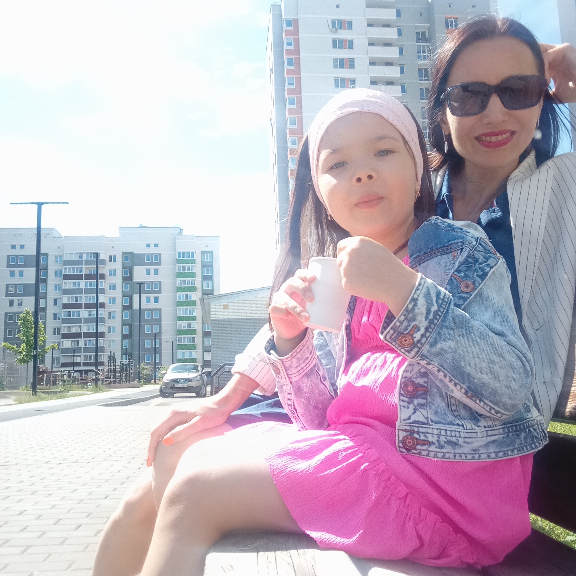 An adult woman and a young girl are seated on an outdoor bench in a residential area of Izhevsk, Russia, on a sunny day. The woman, positioned on the right, has dark hair, wears dark sunglasses, a blue denim shirt, and a white pinstriped blazer. She has red lipstick and pearl earrings, and a slight smile, with her left hand behind her head. The girl, on the left, wears a bright pink dress, a light blue denim jacket, and a light pink headband. She holds a white cup with both hands, and her fingernails are painted orange. They are sitting on a light-colored bench. In the background, modern multi-story apartment buildings with grey, white, and accent-colored facades (green, brown, orange) are visible under a bright sky with sparse clouds. A paved street runs alongside the bench, with a light-colored sedan (Russian license plate "862 HKO 18", indicating the Udmurt Republic where Izhevsk is located) parked near the curb. Several black, T-shaped streetlights line the street, and playground structures are partially visible behind them. The foreground ground surface is composed of interlocking pavers.