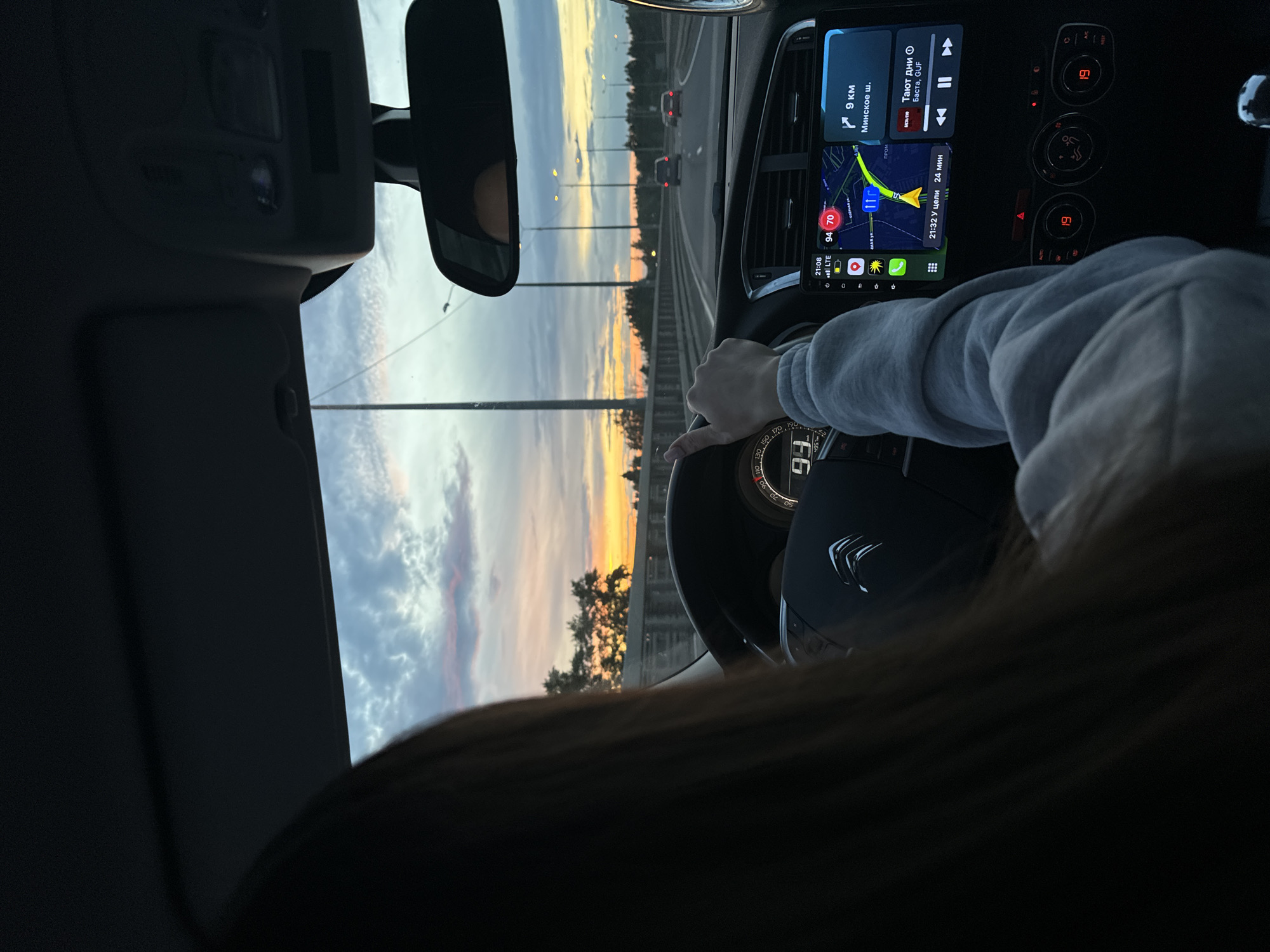 A view from inside a car shows a driver on a highway, heading west, possibly near Selyatino, Russia based on the navigation display that reads “9 km to Minskoye Shosse." The dashboard displays a navigation app, playing music by Basta & GUF. It's dusk or dawn, with a colorful sky visible through the windshield, with the speed showing as 99. Other cars are on the road ahead. The driver's hand is visible on the steering wheel, along with part of their hair.
