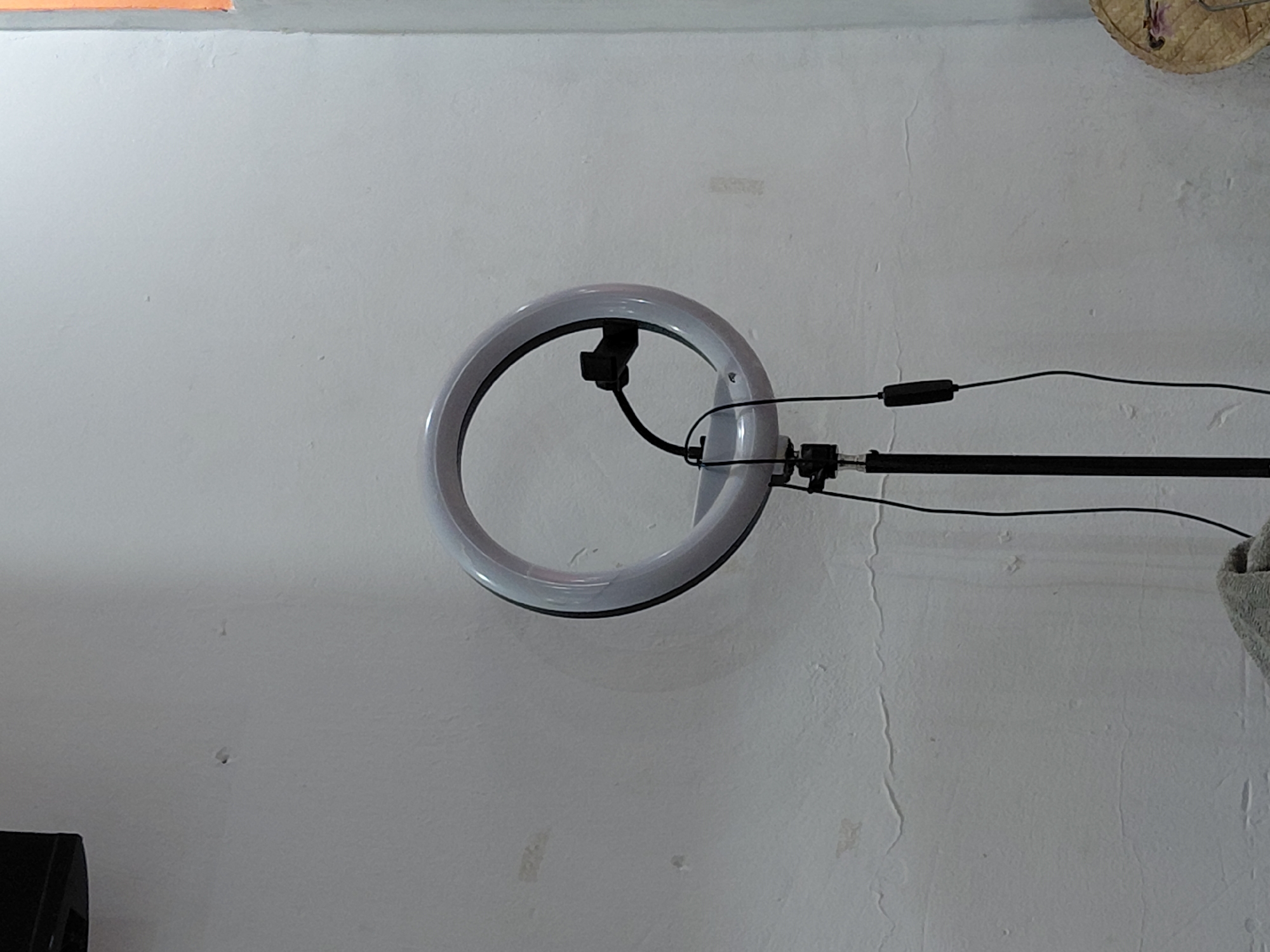 The image captures an indoor scene dominated by a white ring light, which appears to be illuminated with a soft glow, suggesting it is either on or reflecting ambient light. The ring light is mounted on a black stand or boom arm, positioned against a light-colored, textured wall. Wires extend from the ring light along the stand, incorporating a rectangular control component, likely a power switch or dimmer.
The wall, which occupies most of the background, is light-toned, possibly white or off-white, and shows signs of wear, including visible cracks, minor discolorations, and an uneven texture. In the upper left corner, a small section of an orange surface is visible, possibly part of an adjacent wall or ceiling. The upper right corner reveals a portion of a woven object, resembling straw or wicker, which could be a decorative item or part of a fixture. At the bottom left, a dark, angular object, possibly a speaker or piece of equipment, is partially visible. Further to the right, a gray, textured object, appearing to be a folded cloth or towel, is also partially cropped.
The arrangement of the ring light and stand indicates a setup for photography or video recording. No people are present, and no specific activity is explicitly shown beyond the static presence of the equipment. The visible details suggest a functional space, perhaps a home studio or workshop, located in Santa Ana, Venezuela. There is no visible text in the image.