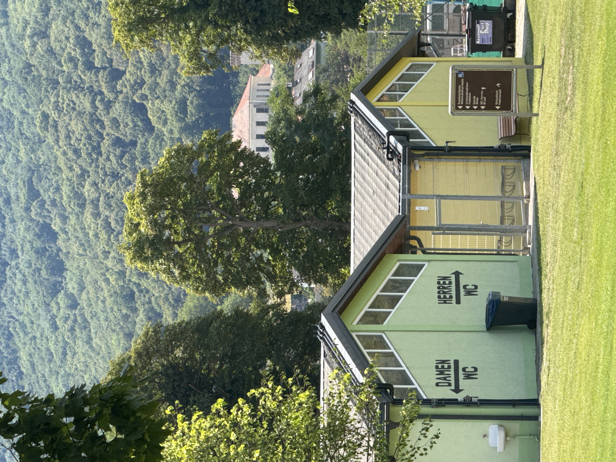 The image depicts a public restroom facility with separate entrances for "Damen" (Women) and "Herren" (Men), indicated by directional arrows. The buildings are painted in vibrant green and yellow. To the left of the "Damen" entrance, a white sink is mounted on the wall. A dark trash bin is situated near the "Herren" entrance. In front of the buildings, a manicured green lawn is visible. A brown signpost with multiple informational panels stands to the right, listing amenities such as "Umkleidekabinen" (changing rooms) and "WC Herren" (Men's WC). Behind the restrooms, a chain-link fence and more trash receptacles are present. In the background, lush, green forested hills rise, characteristic of the landscape surrounding Hainburg an der Donau, Austria, with some buildings visible amongst the trees and on the hillside. The scene is bathed in sunlight, suggesting a daytime setting.