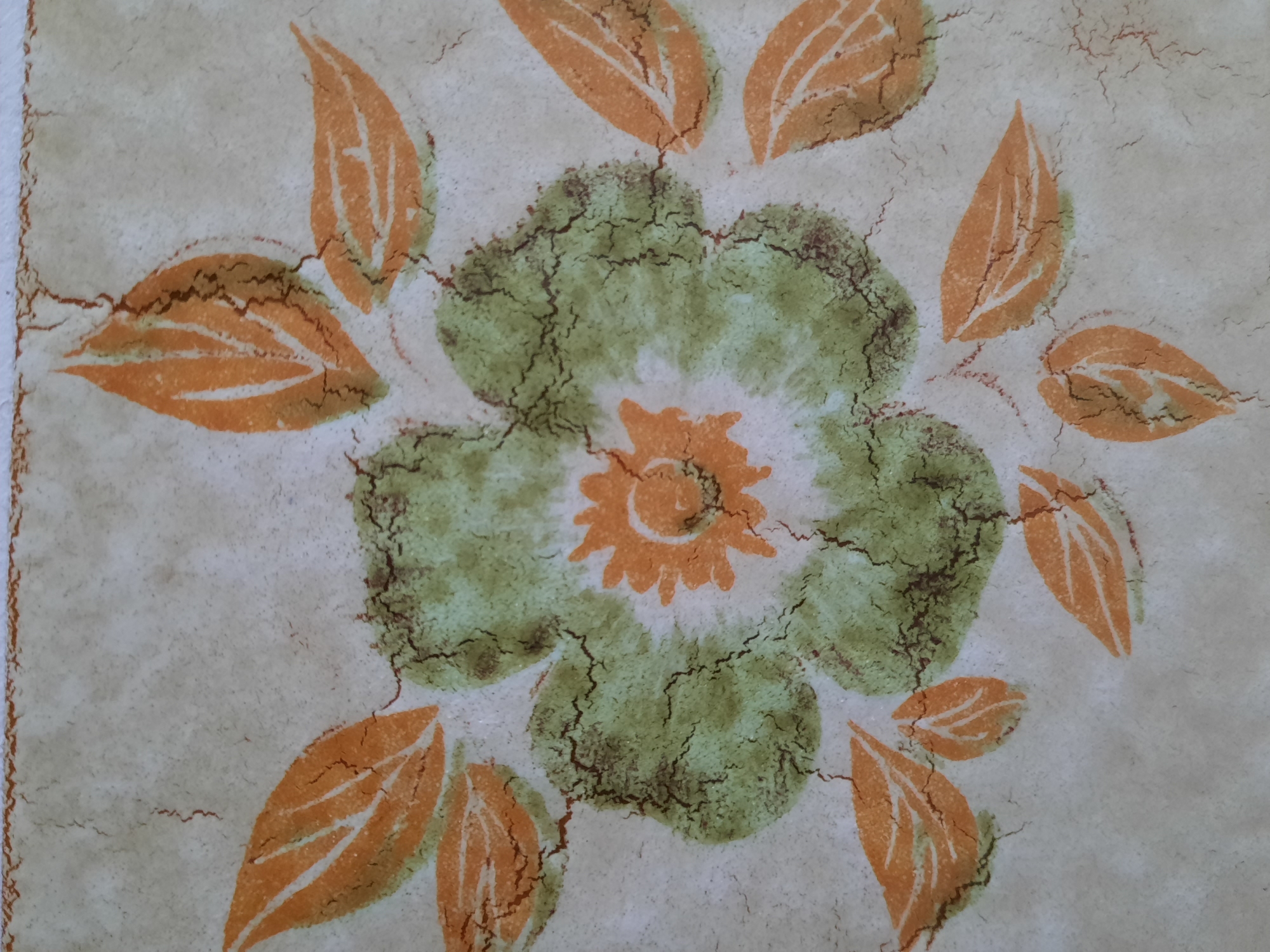 This is a close-up, overhead photograph of a vintage, possibly ceramic or laminate, surface featuring a floral pattern. The dominant colors are a pale beige background, green for the flower's petals, and orange for the flower's center and surrounding leaves. The design shows a stylized flower with a central orange sun-like element surrounded by textured green petals. Several orange leaves are arranged radially around the flower. The entire surface is covered in fine, dark crackling, resembling crazing on pottery or a distressed finish. There are no people, objects, or environmental elements that suggest a specific location within the city of Rosenheim, Germany. The image focuses solely on the decorative pattern of the surface.