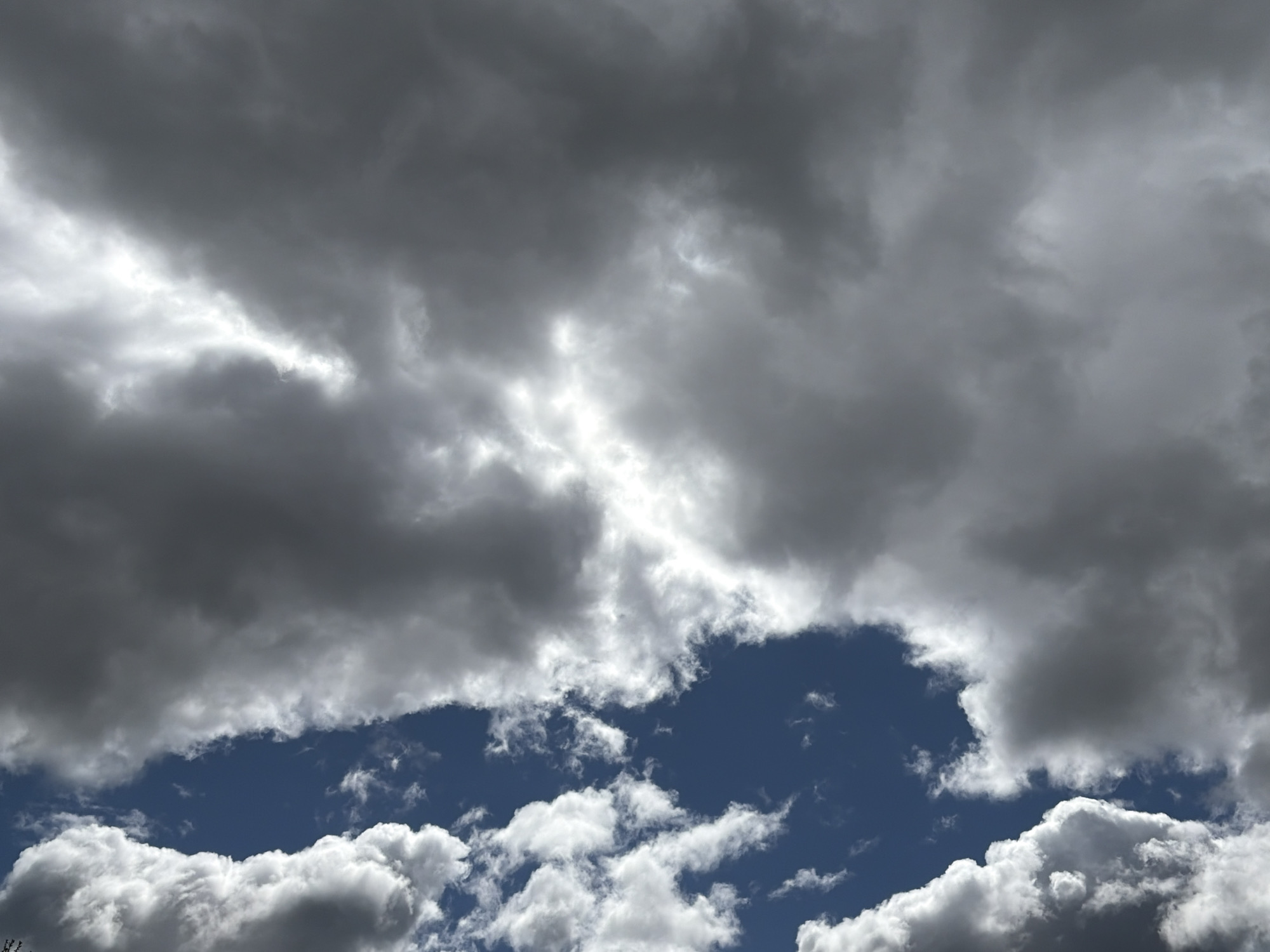 The image shows a cloudy sky. The upper portion of the image is dominated by dark, overcast clouds, while a patch of blue sky is visible in the lower center, framed by lighter, white clouds. The location is unknown.